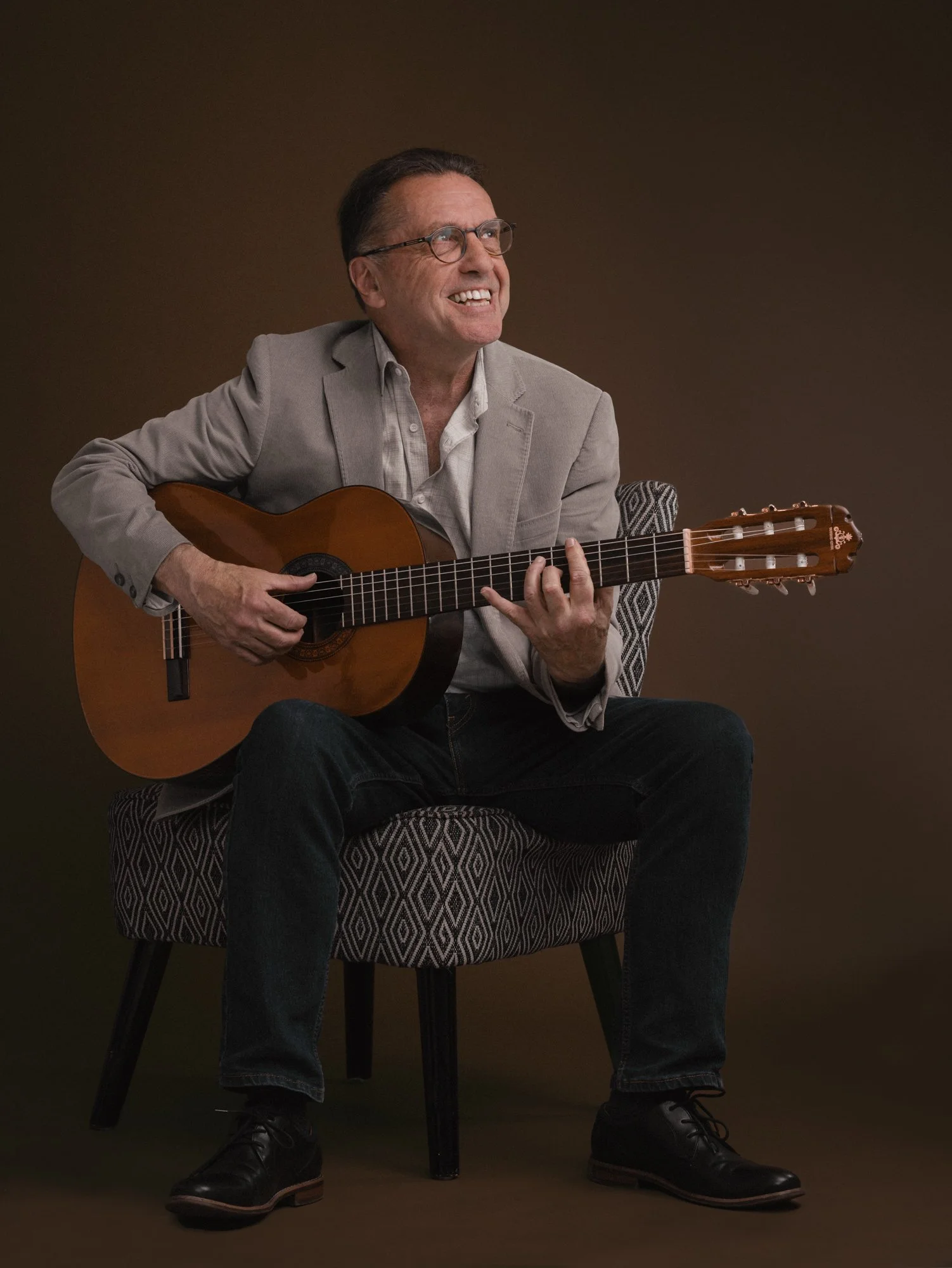 A man sitting on a patterned chair, playing an acoustic guitar, wearing glasses, a light blazer, a white shirt, dark jeans, and black shoes, with a brown background.