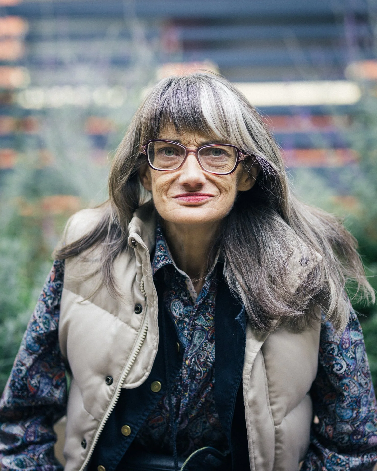 A woman with long gray hair and glasses, wearing a beige vest, a patterned shirt, and a dark blazer, outdoors with a blurred background of buildings and trees.