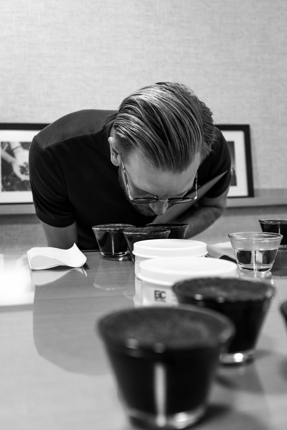 A man with glasses and slicked-back hair is smelling a glass cup on a table with multiple cups filled with dark liquid, in a room with framed photos on the wall.