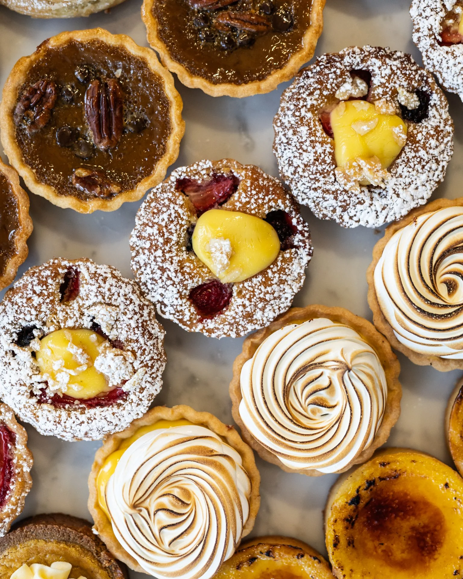 Variety of mini tarts with different fillings and toppings, including chocolate, caramel, fruit, and meringue, arranged on a tray.