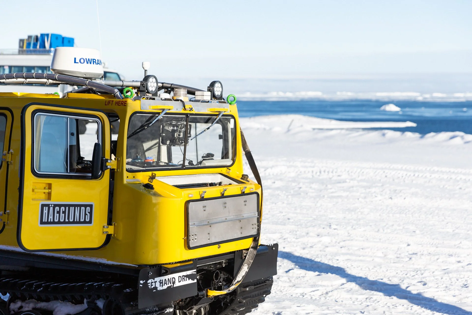 A yellow snow grooming machine with tracks, labeled 'HÄGGLUNDS,' operating on snow at a snowy landscape with a body of water and ice in the background.