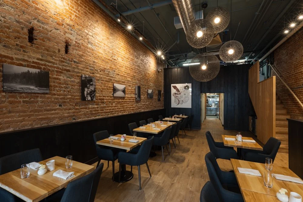 Interior of a modern restaurant with exposed brick walls, wooden floors, black chairs, and tables set with glasses, napkins, and salt shakers. Hanging spherical light fixtures are on the ceiling, and black and white artworks are on the wall.