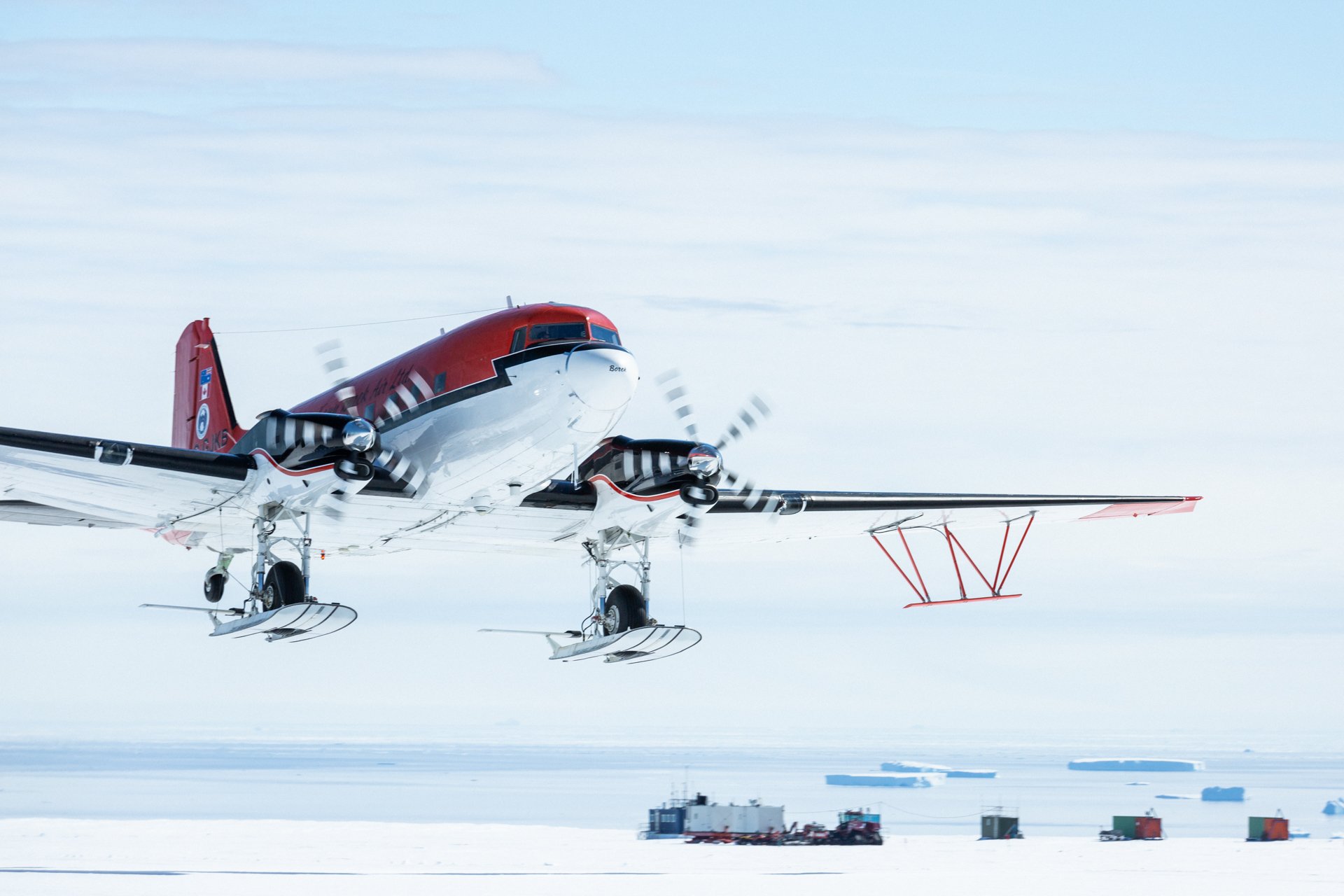 Propeller-driven airplane landing on snow-covered surface with support structures and containers in background