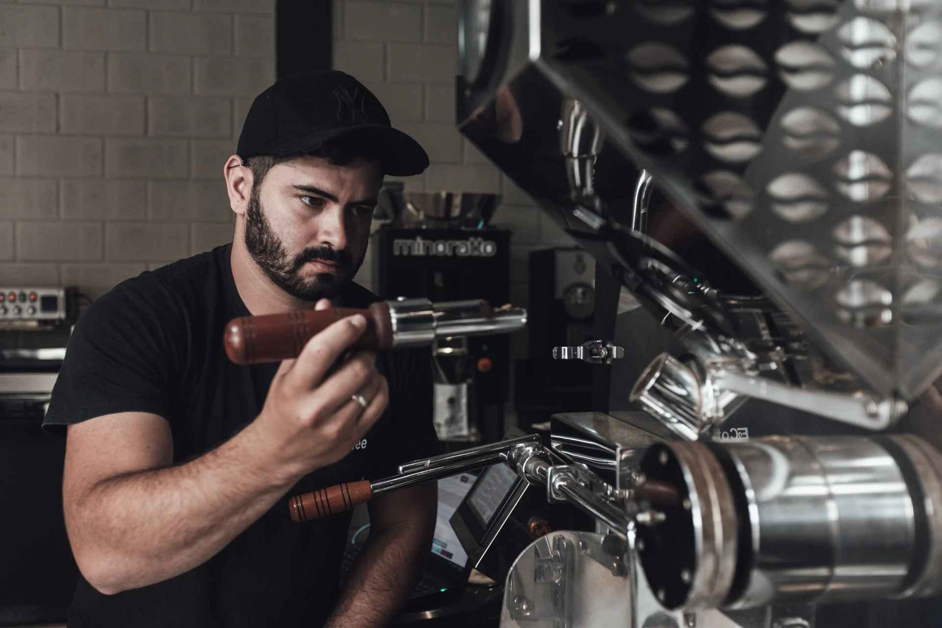 A man with a beard and black cap making a latte art coffee in a commercial espresso machine in a coffee shop.