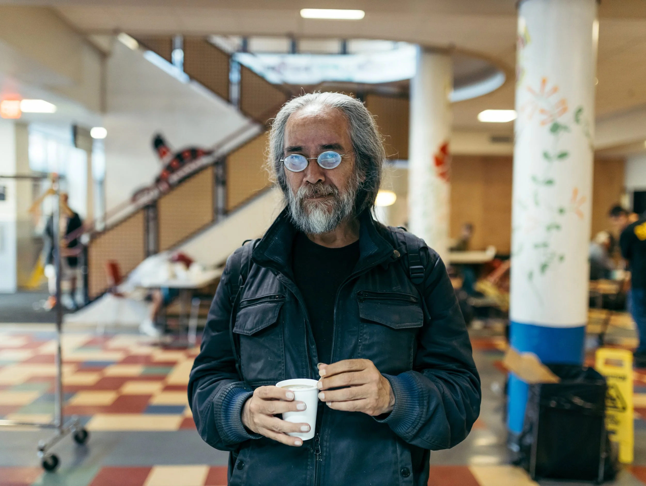 An older man with a beard and glasses, wearing a black jacket, holding a cup of coffee or hot beverage inside a brightly lit indoor space, possibly a community center or cafeteria.