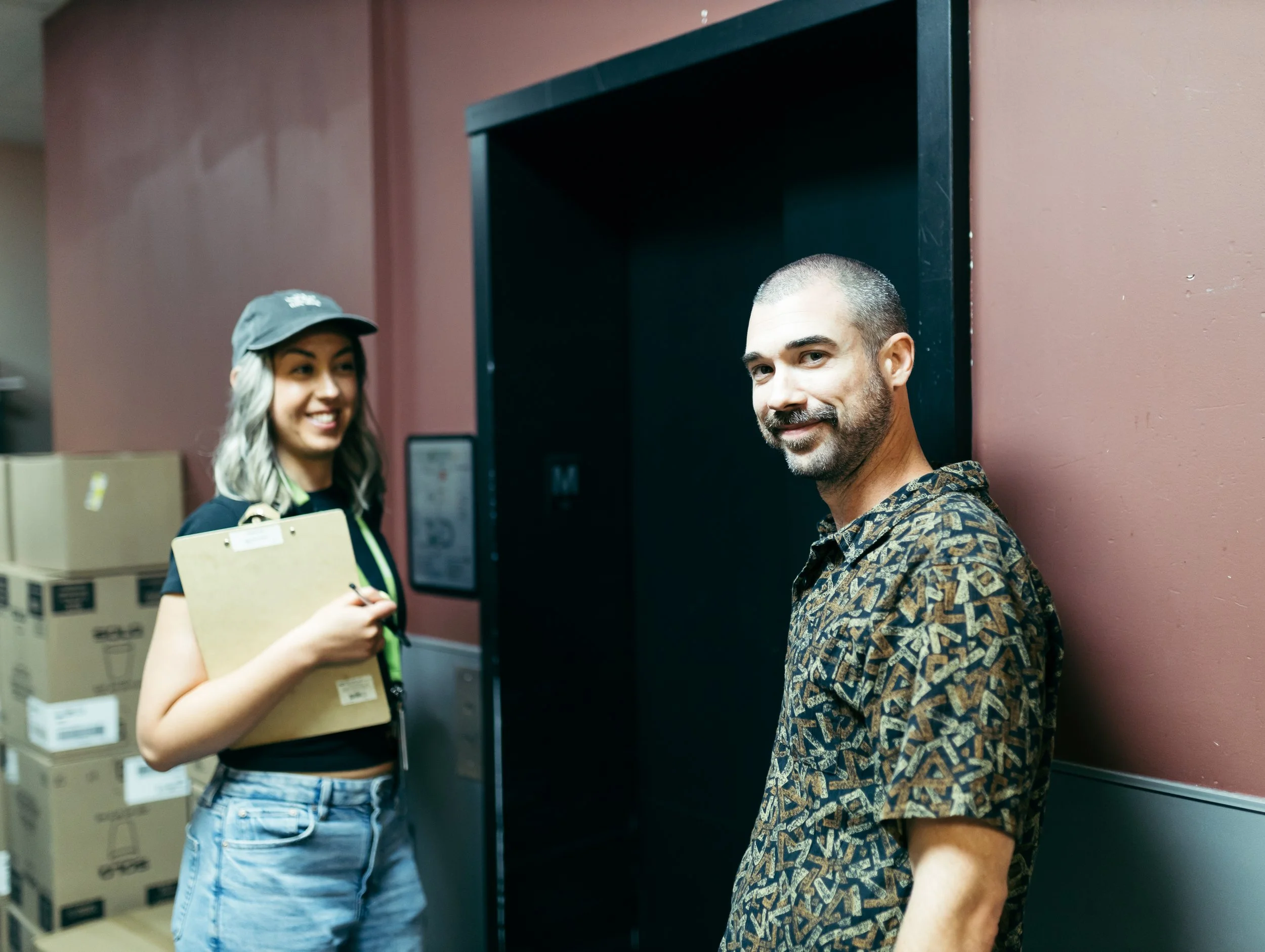 A smiling man with a beard and short hair is standing in front of an elevator, with a woman holding a clipboard and wearing a cap and casual clothes standing nearby, surrounded by stacked boxes in an indoor setting.