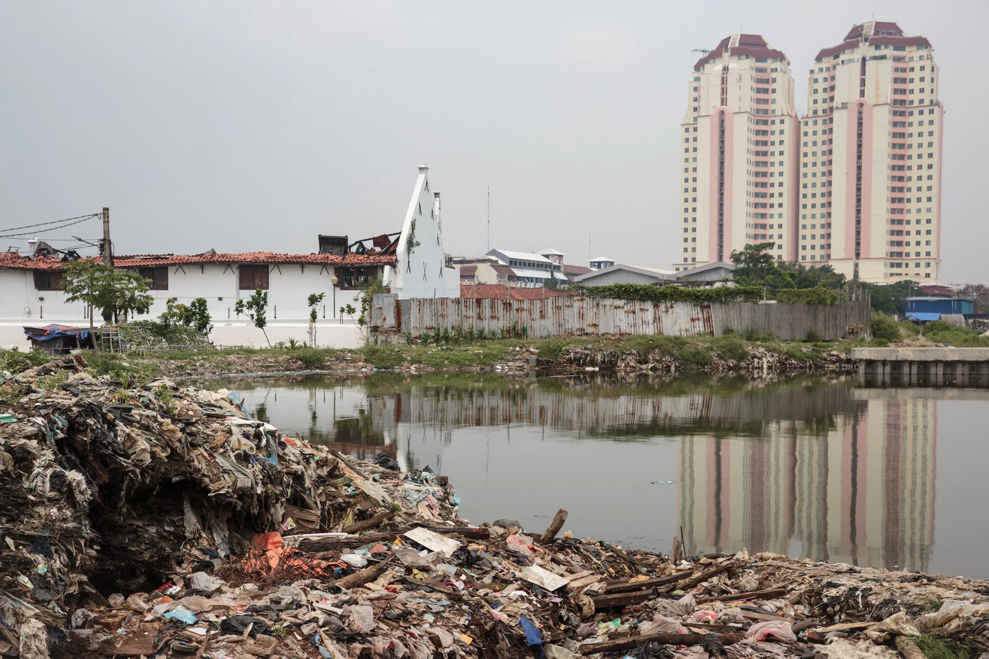 A riverbank cluttered with trash and debris in the foreground, with a body of water reflecting nearby buildings. In the background, there are small, older houses with red tiled roofs, and two tall, modern high-rise buildings with a cloudy sky above.