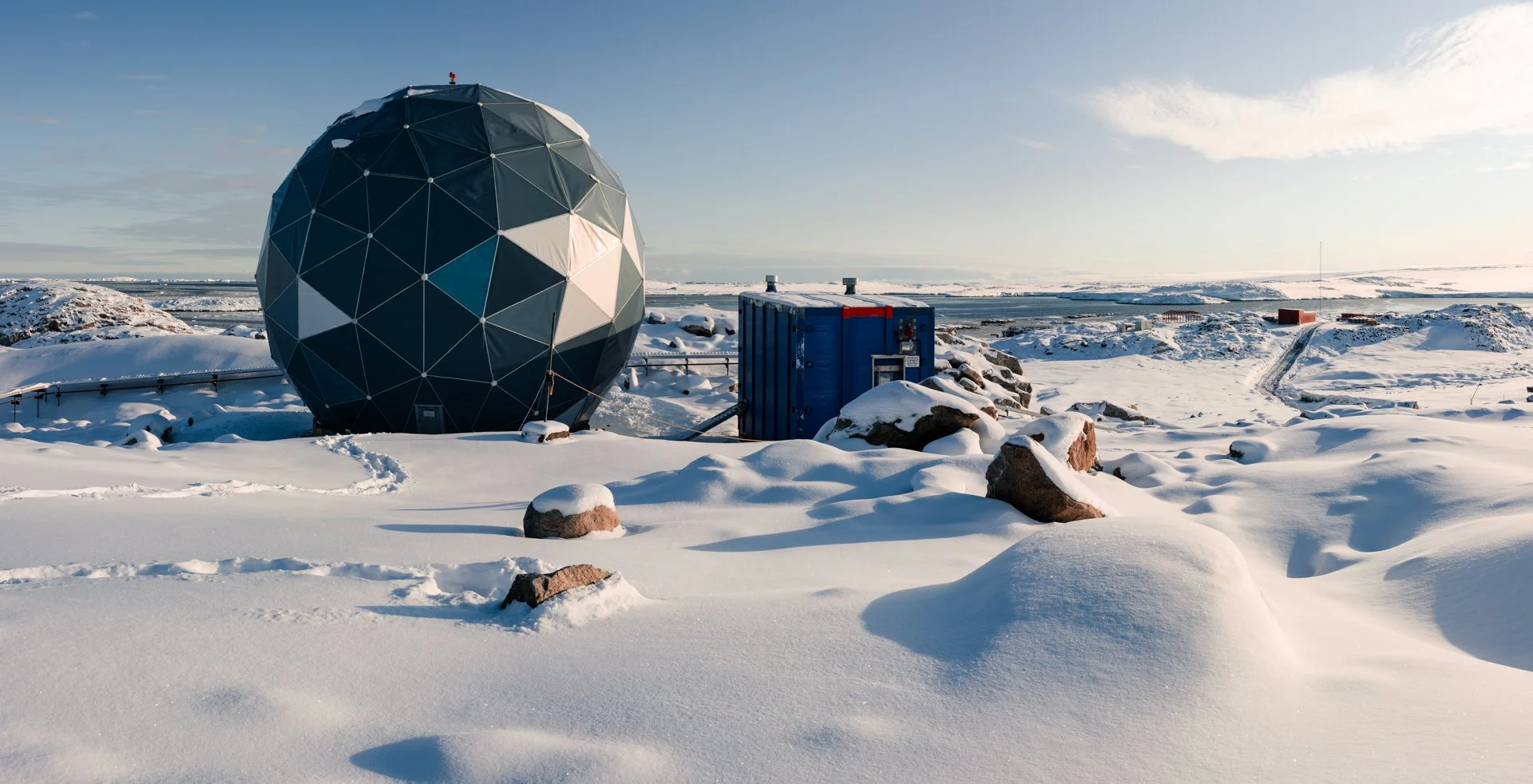 A snow-covered landscape with a geodesic dome and a blue container surrounded by rocks, with footprints and a few distant buildings under a clear sky.