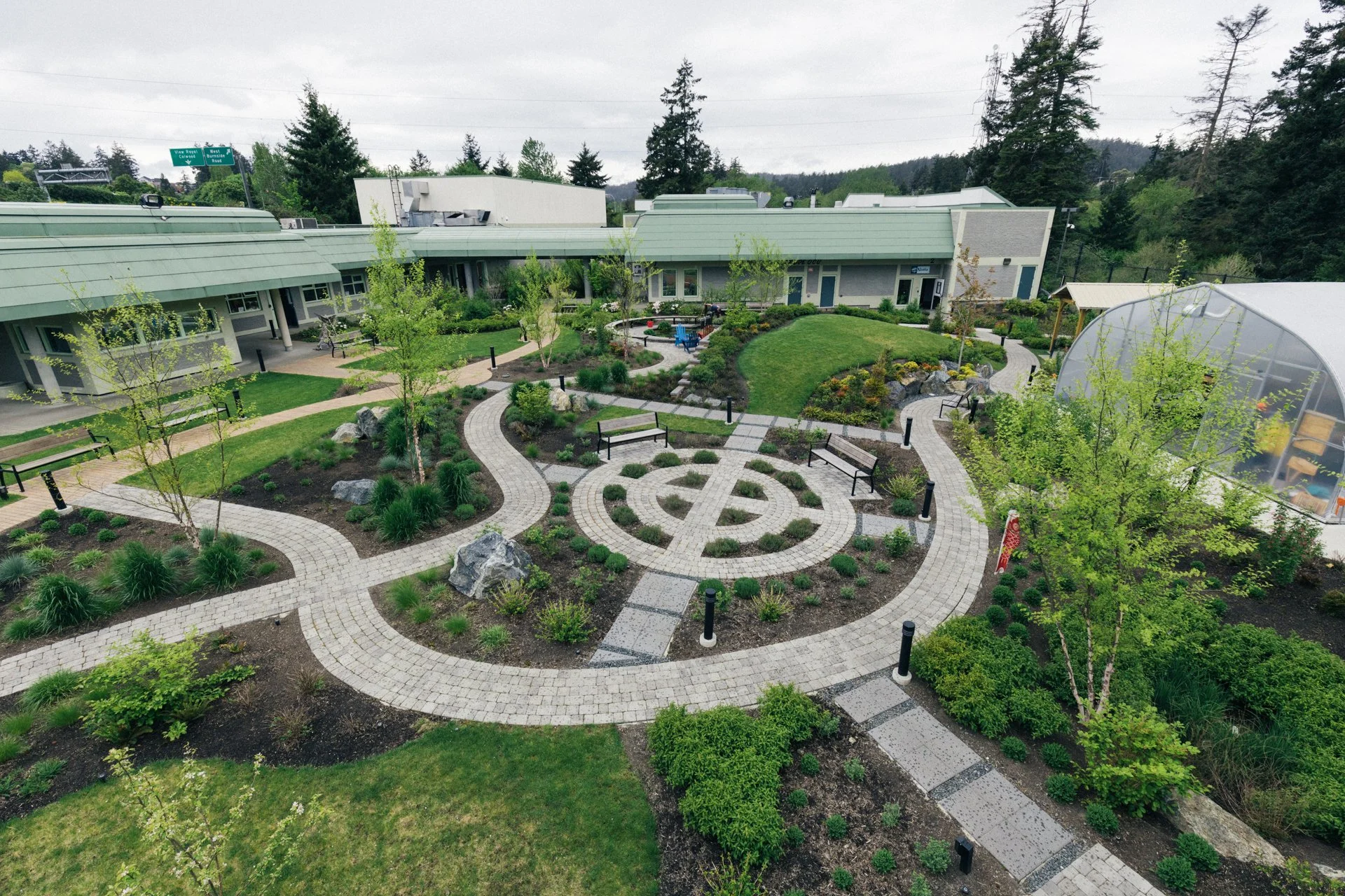 A courtyard garden with winding brick pathways, green grass, small shrubs, and young trees, surrounded by modern buildings with green roofs and large windows, under an overcast sky.