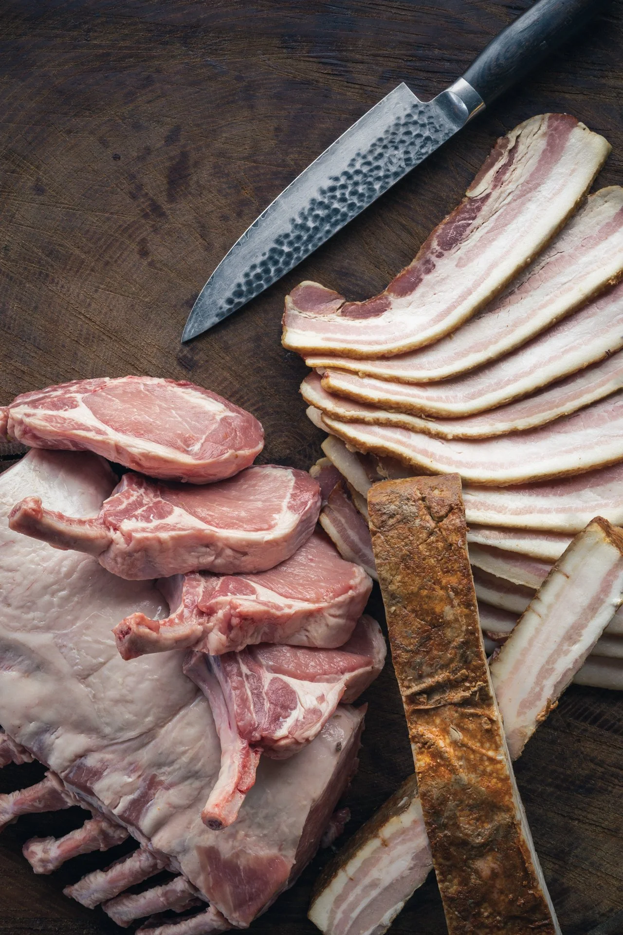 A cutting board with slices of bacon, pork chops, and ribs, a sharp knife, and two seasoned or cooked rib pieces.