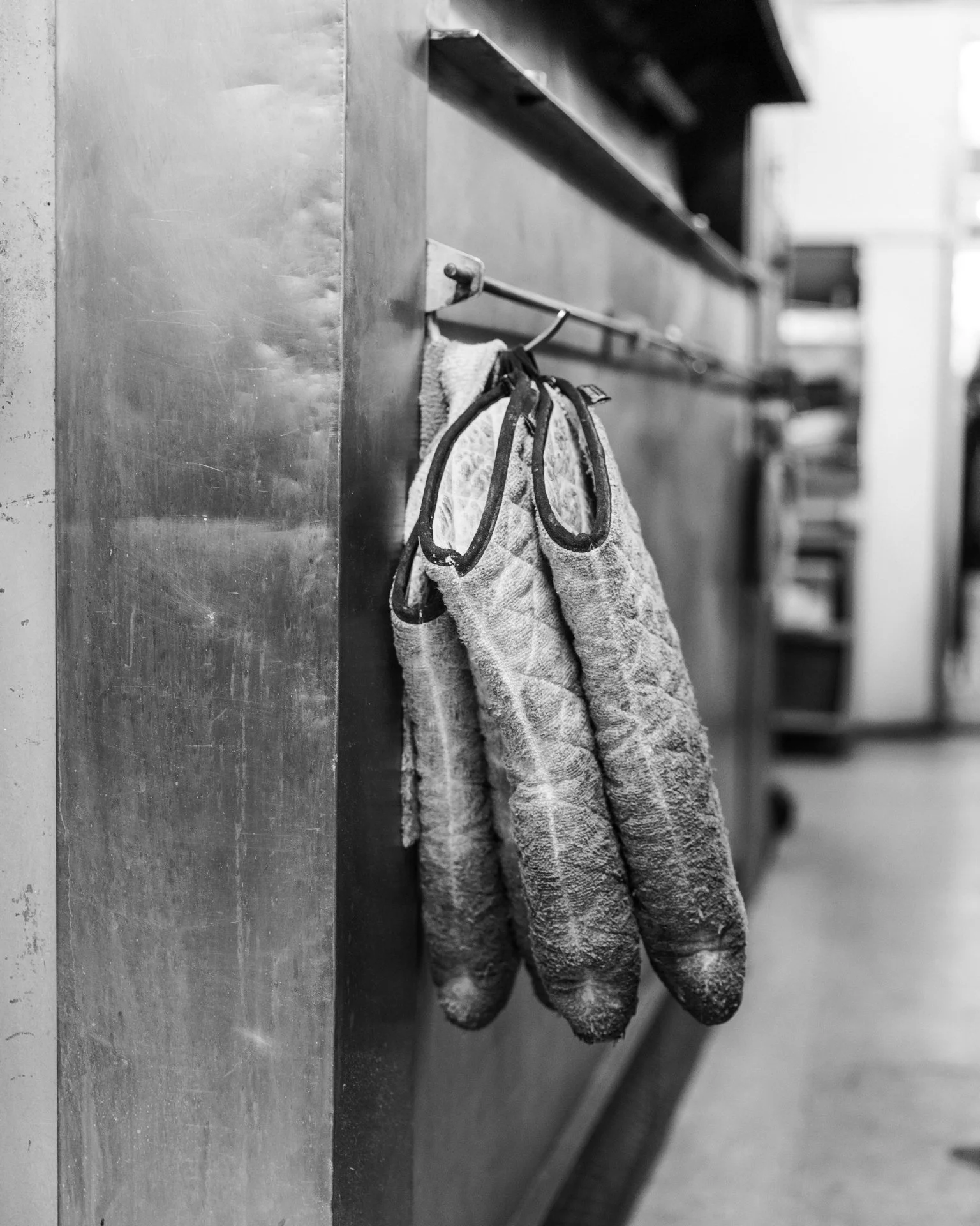 Black and white photo of a pair of hanging work gloves and a cloth attached to a hook on a wall in what appears to be a workshop or industrial setting.