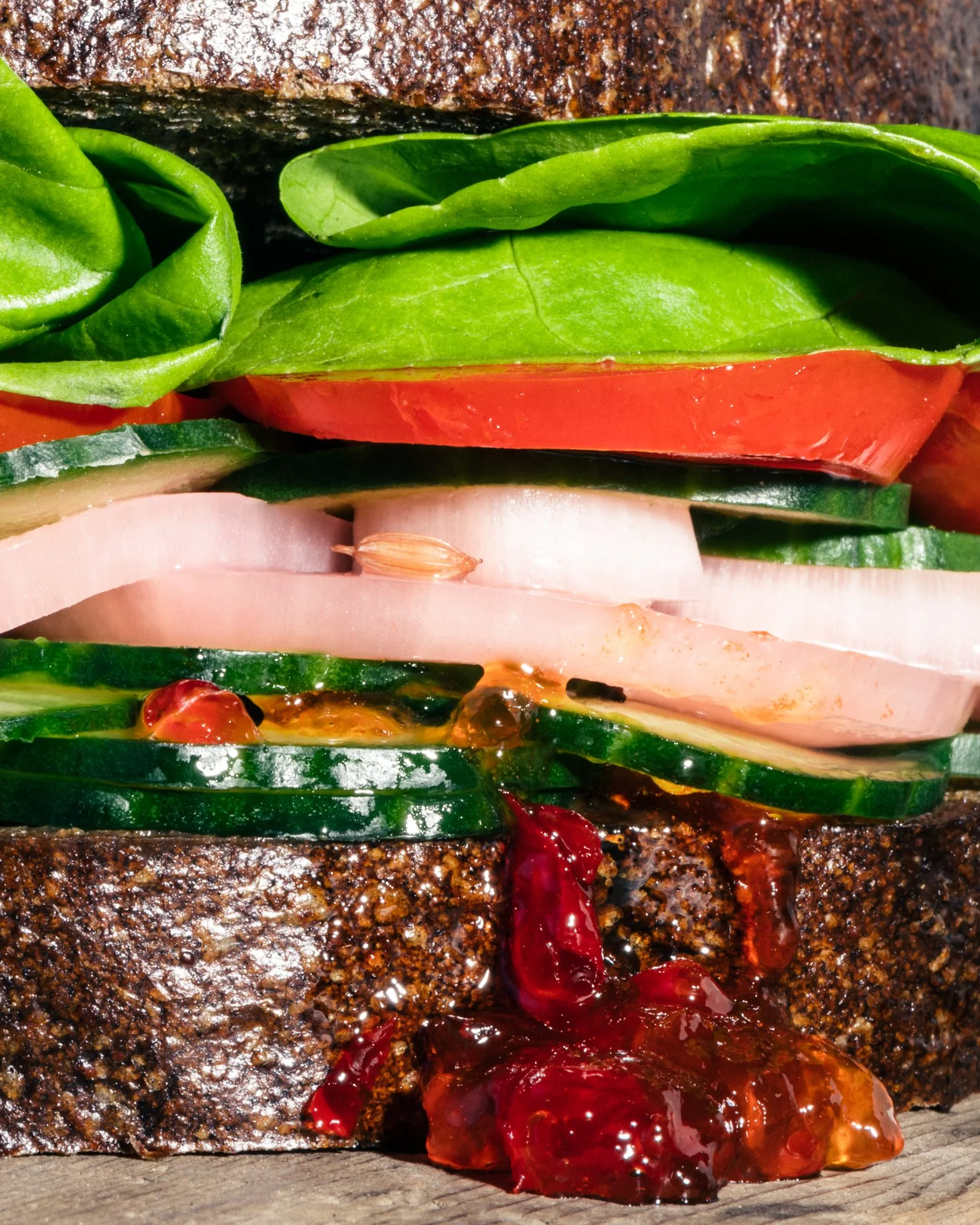 Close-up of a grilled burger patty with fresh vegetables, including slices of tomato, cucumber, onion, spinach, and red pepper, with ketchup on a wooden surface.