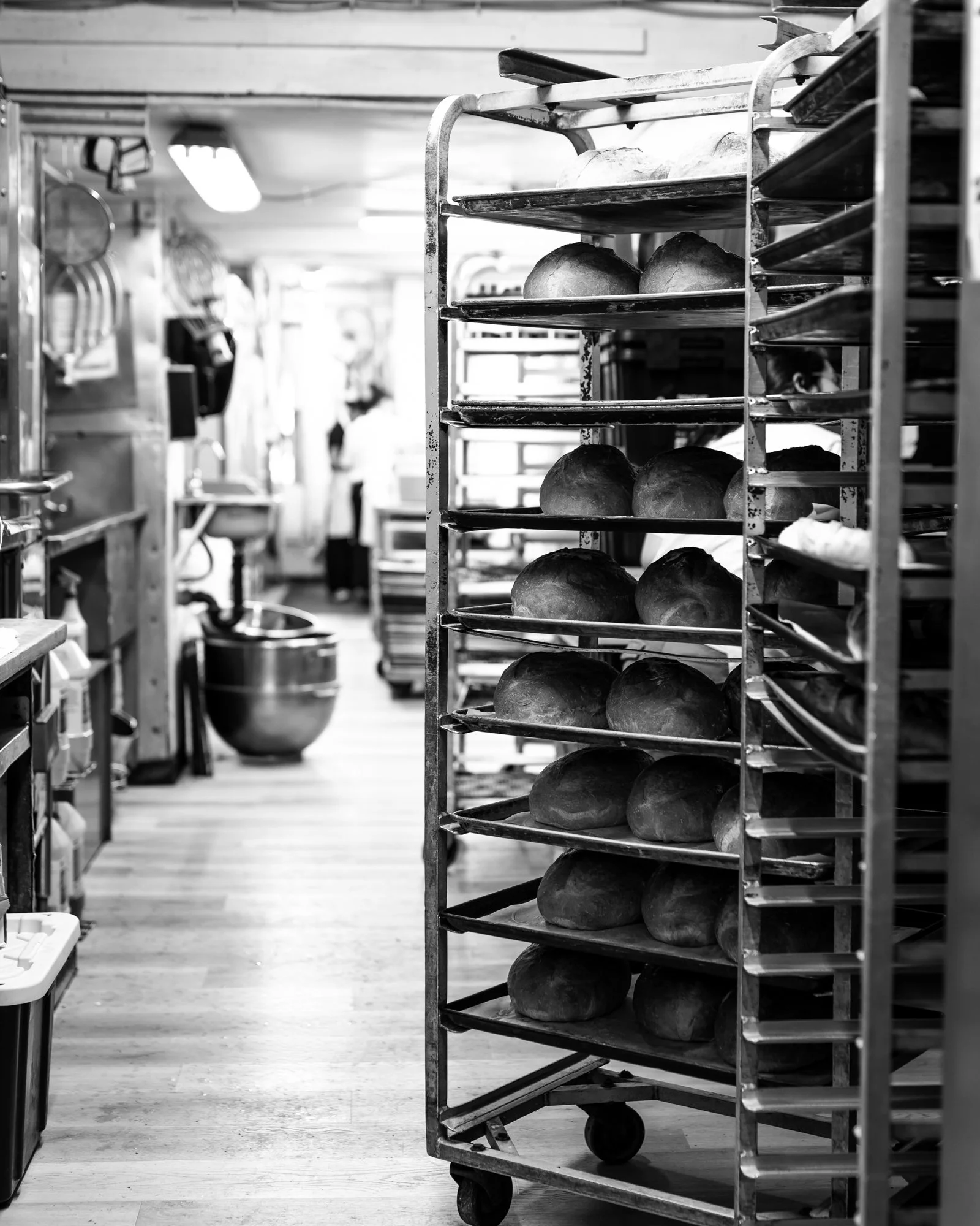 Black and white photo of a bakery kitchen with a rack of freshly baked bread in the foreground and cooking utensils hanging on the wall in the background.