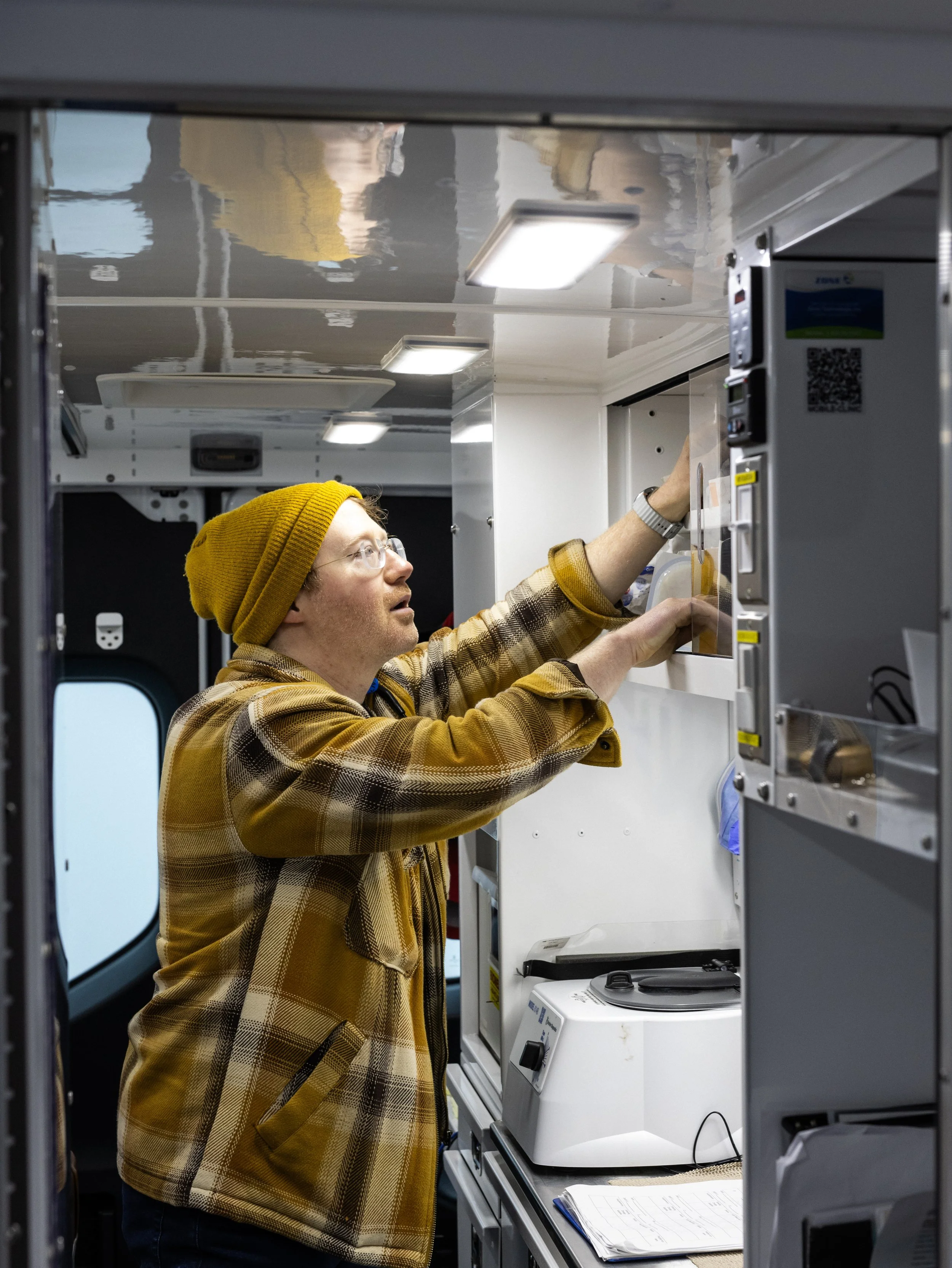 A person in a yellow beanie and plaid jacket working on electronic equipment inside a mobile lab or medical van.