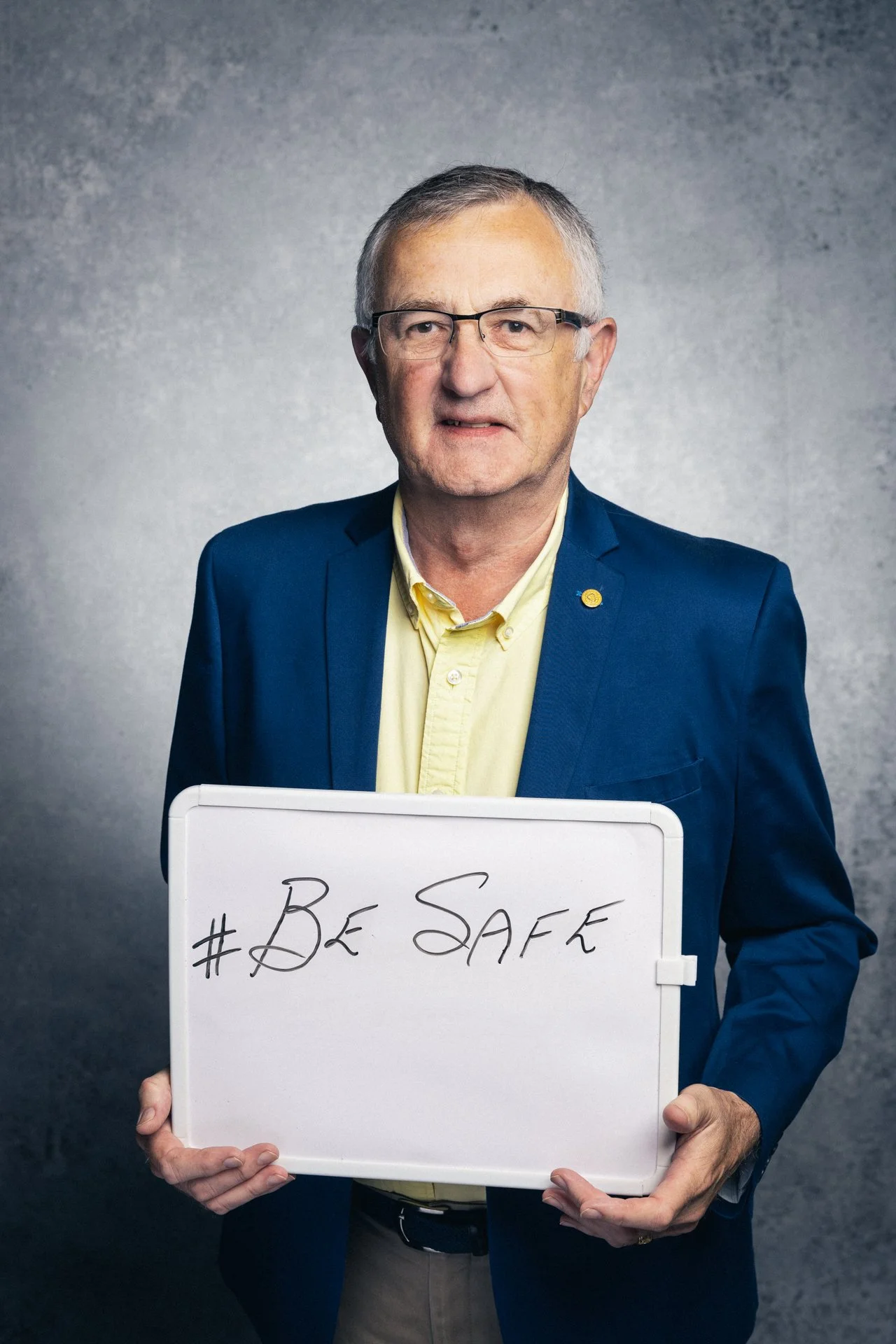 An older man with glasses, wearing a yellow shirt and a blue blazer, holding a whiteboard that reads '#BeSafe'.