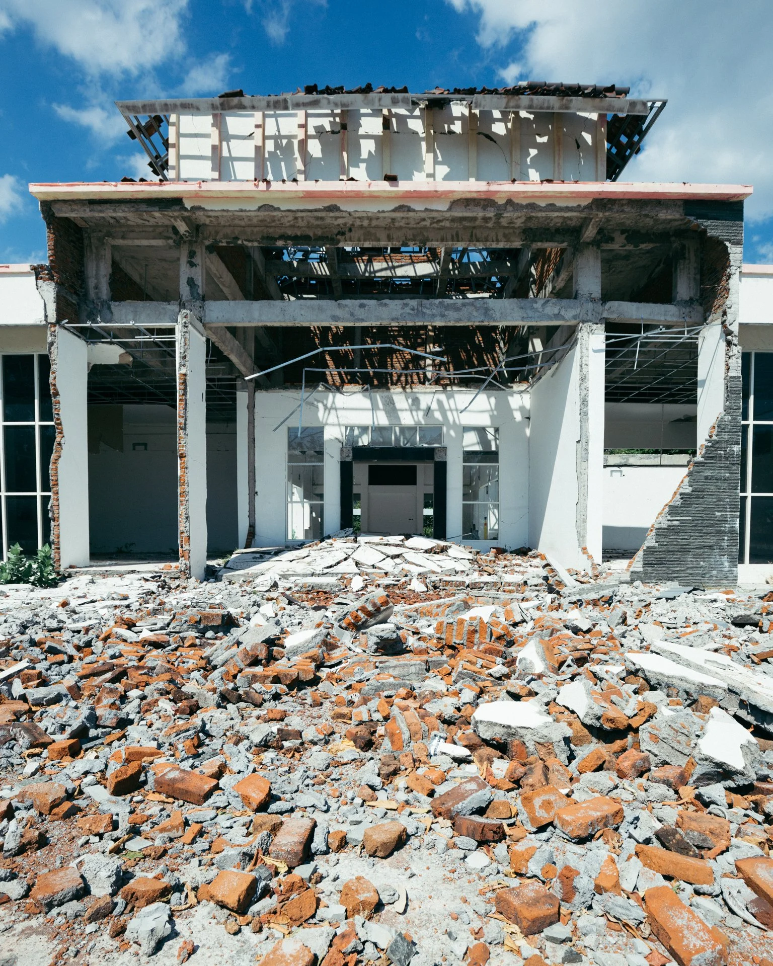A partially demolished building with the front wall removed, exposing the inside structure, debris and rubble scattered in front, and the sky with clouds in the background.