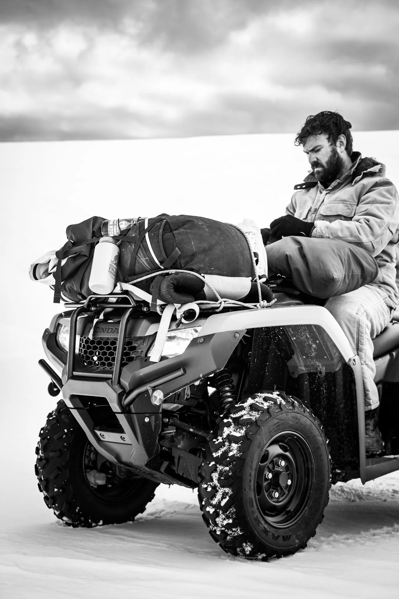 Man riding an all-terrain vehicle (ATV) in a snowy landscape, dressed in winter gear, with a loaded backpack and water bottle tied to the ATV.