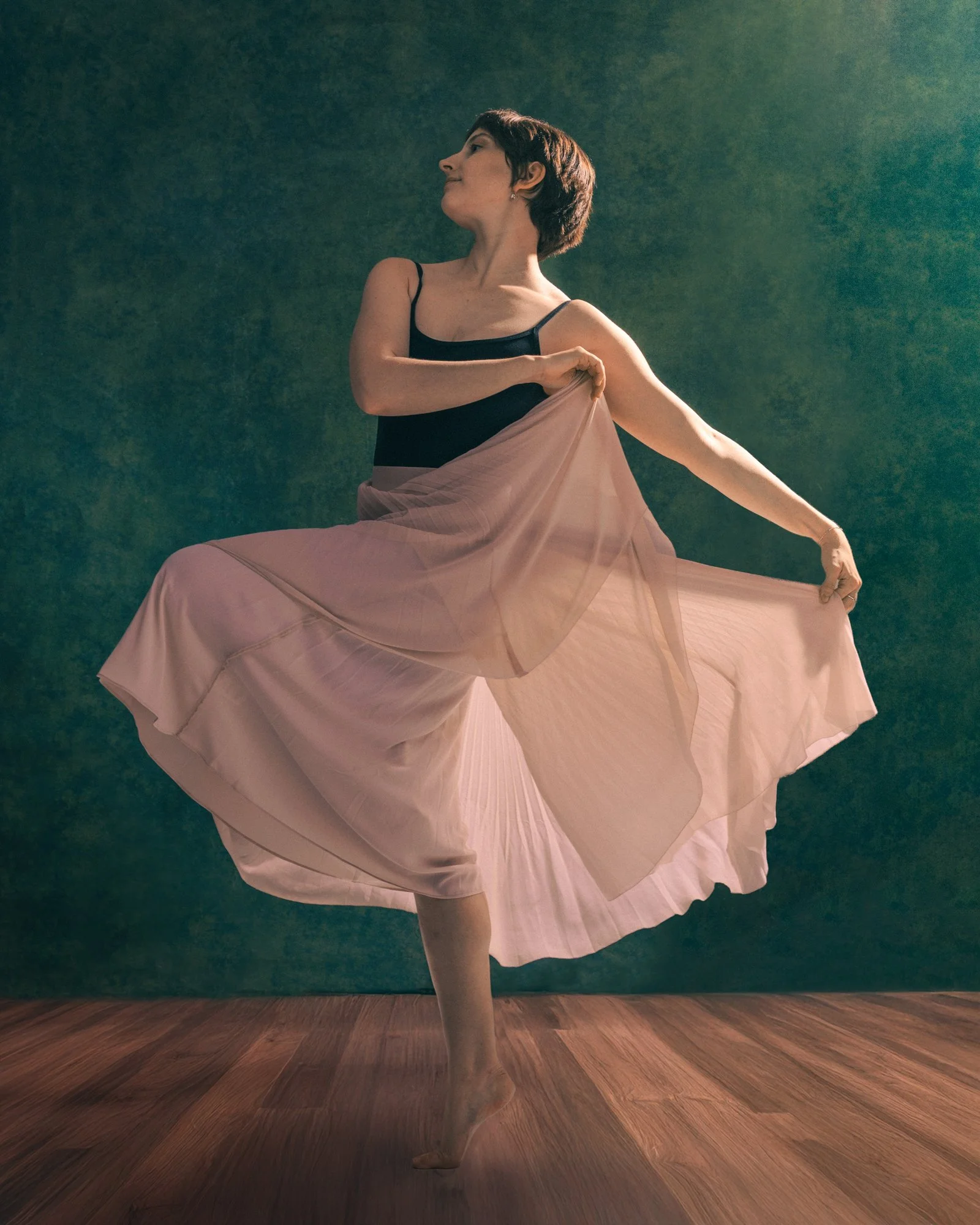 A ballerina in a black leotard and flowing pink skirt dancing on a wooden floor with a green background.