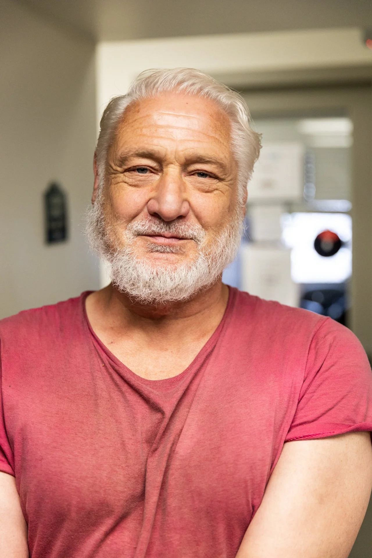 Close-up of an elderly man with a white beard and hair, wearing a pink T-shirt, smiling slightly, standing in a room with a doorway and blurred background.