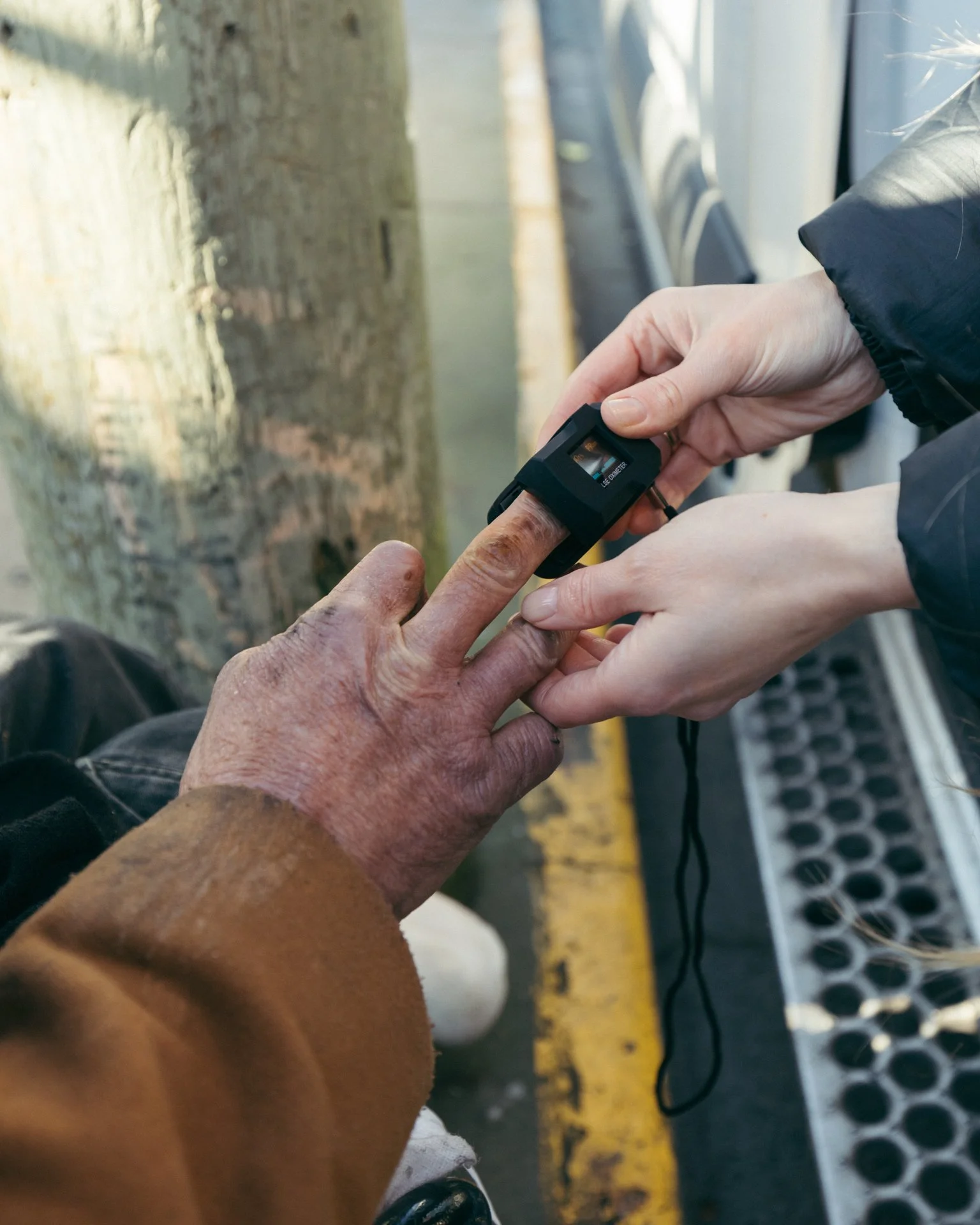 A person with a darker skin tone holds a small digital device, possibly a scanner or meter, near the finger of an older person with lighter skin tone, in what appears to be a transportation setting with a curb and metal grates visible.