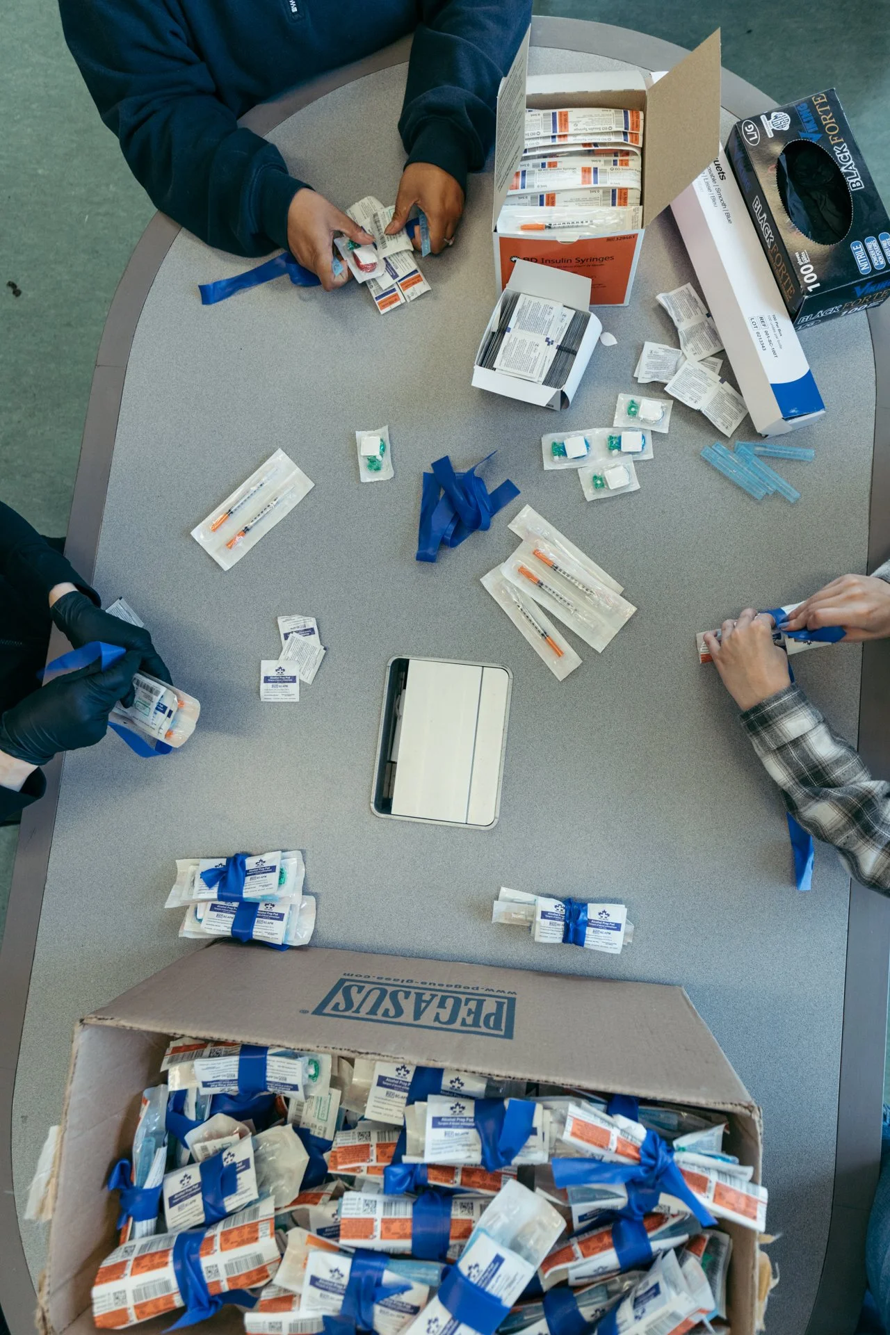 People organizing and packing insulin syringes into boxes, with open boxes of syringes and supplies on a gray table.