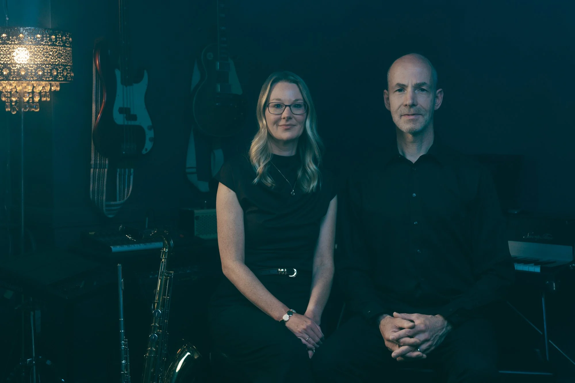 A woman with long blonde hair, glasses, and a black dress sitting next to a man with a shaved head and gray beard, dressed in black, in a music room with guitars hanging on the wall and a keyboard in front of them.