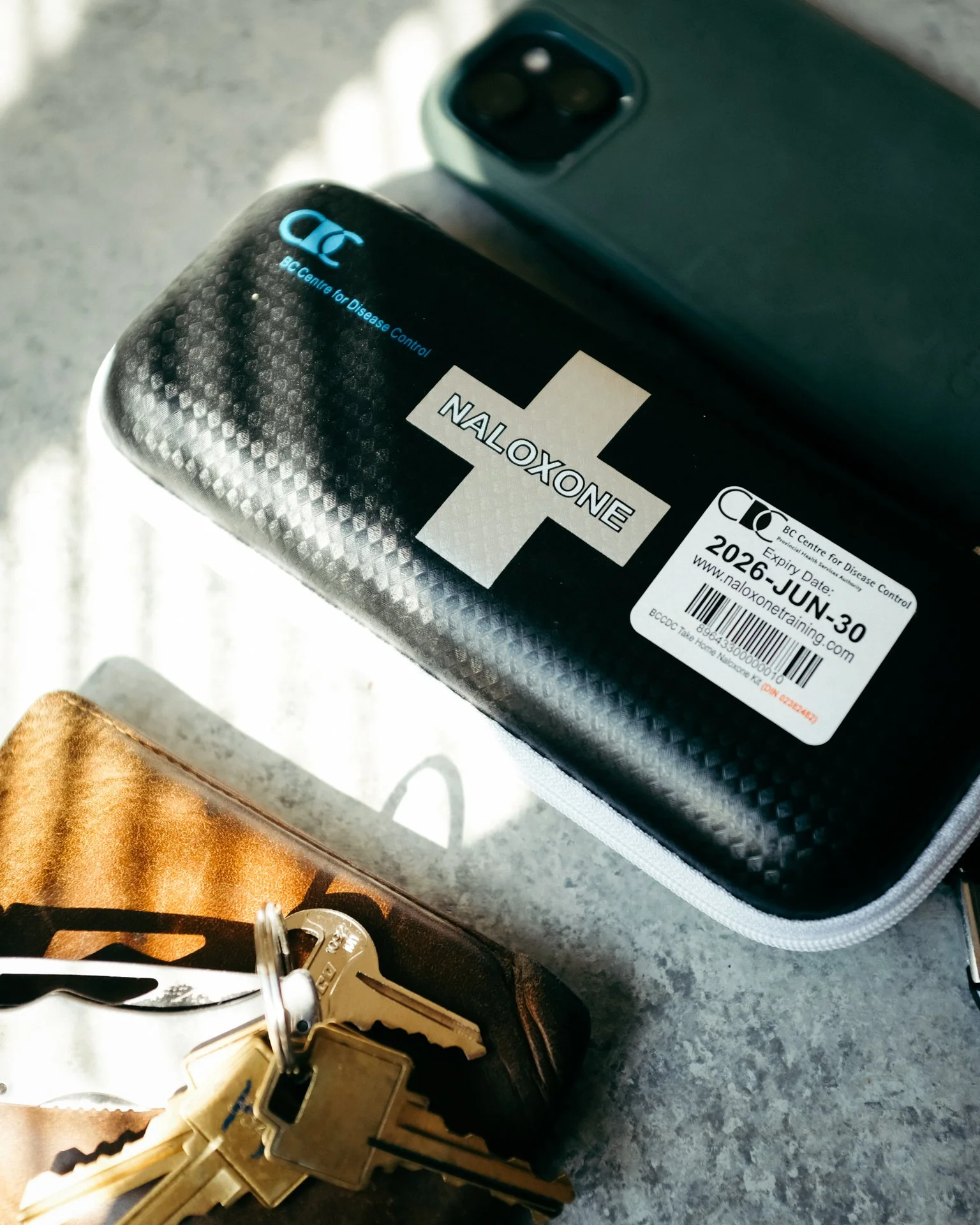 A black electronic device with a cross and blue text reading 'AC Centre for Disease Control'. A white sticker on the device indicates it is a Naloxone kit with an expiration date of June 30, 2026. A set of keys and a golden badge or tag are also visi