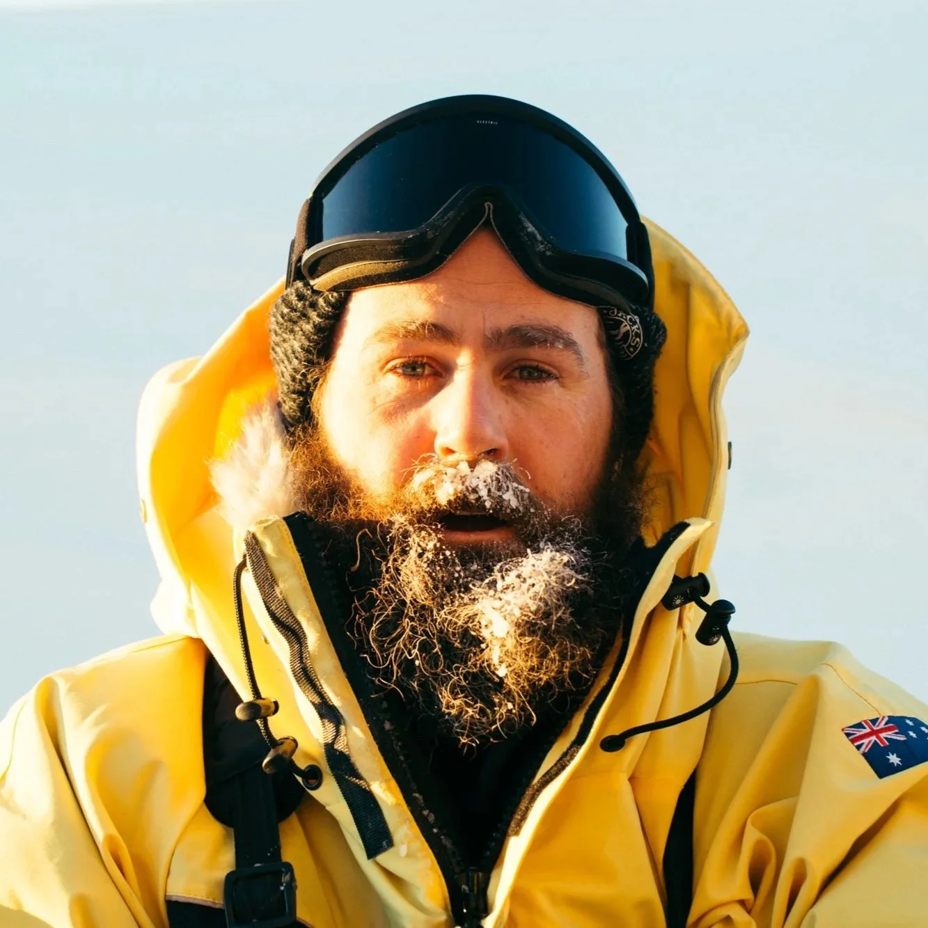 A man with a beard and mustache, wearing a black beanie, ski goggles, and a yellow jacket with a patch of the Australian flag on the sleeve, standing outdoors in cold weather.