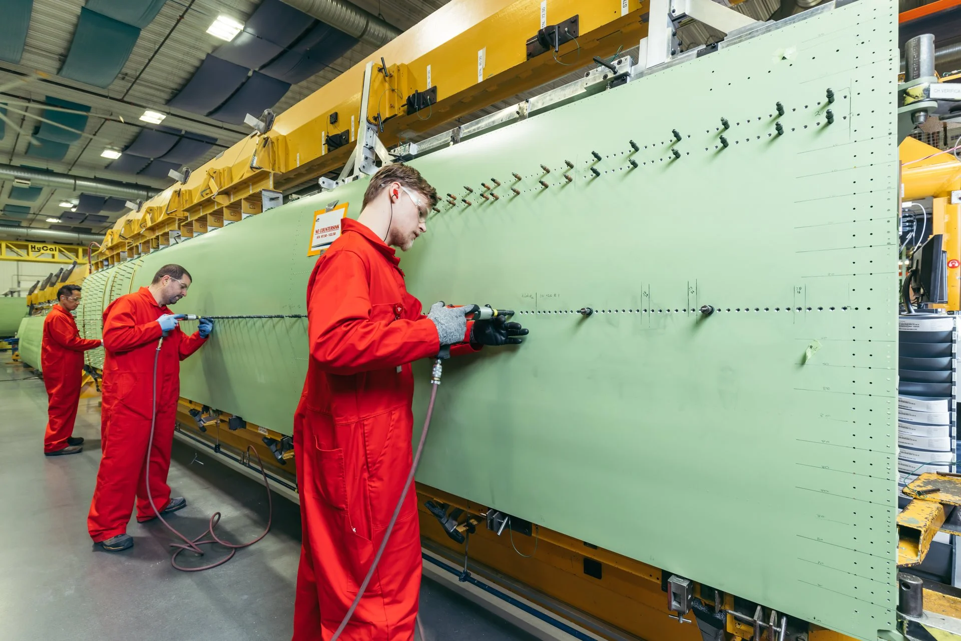 Workers in red coveralls welding and inspecting a large spacecraft or aircraft component inside an industrial facility.