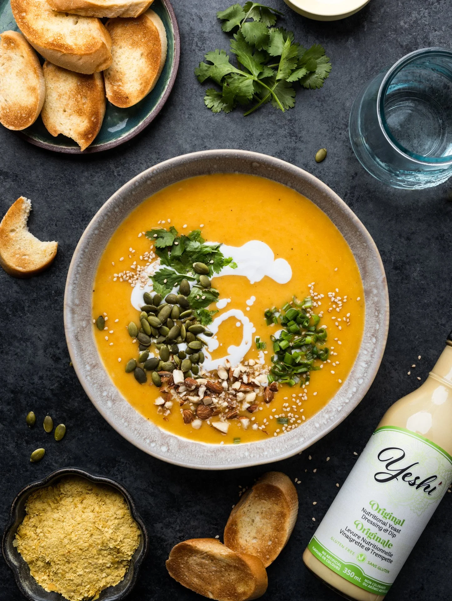Bowl of creamy pumpkin soup topped with cilantro, pumpkin seeds, chopped green onions, sesame seeds, and shredded almonds. Surrounding the bowl are garlic bread slices, a bowl of breadcrumbs, fresh cilantro, a glass of water, a bottle of nutritional 