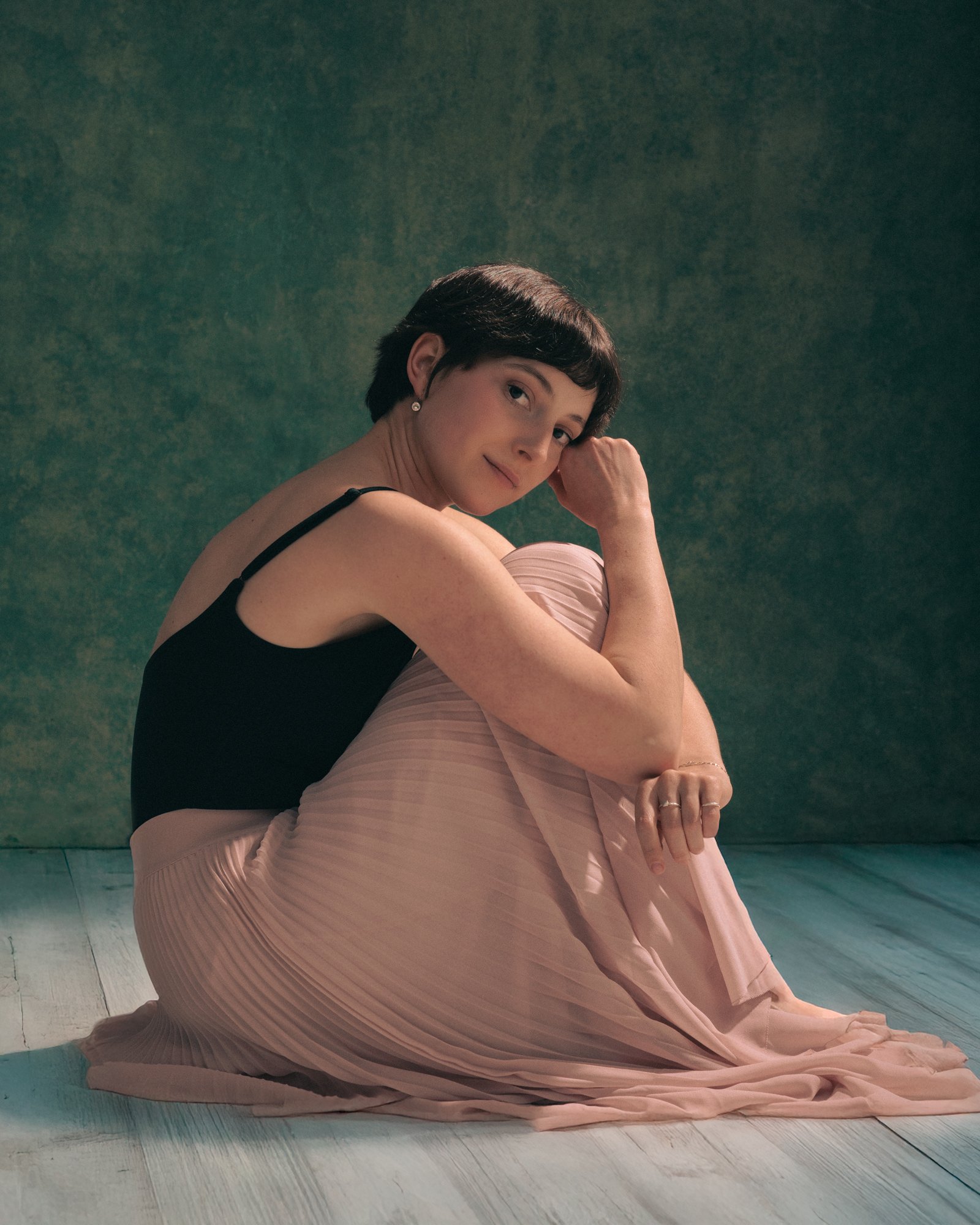 A woman with short dark hair, wearing a black tank top and pink pleated skirt, sitting on the floor against a textured green wall, looking at the camera with a subtle smile.