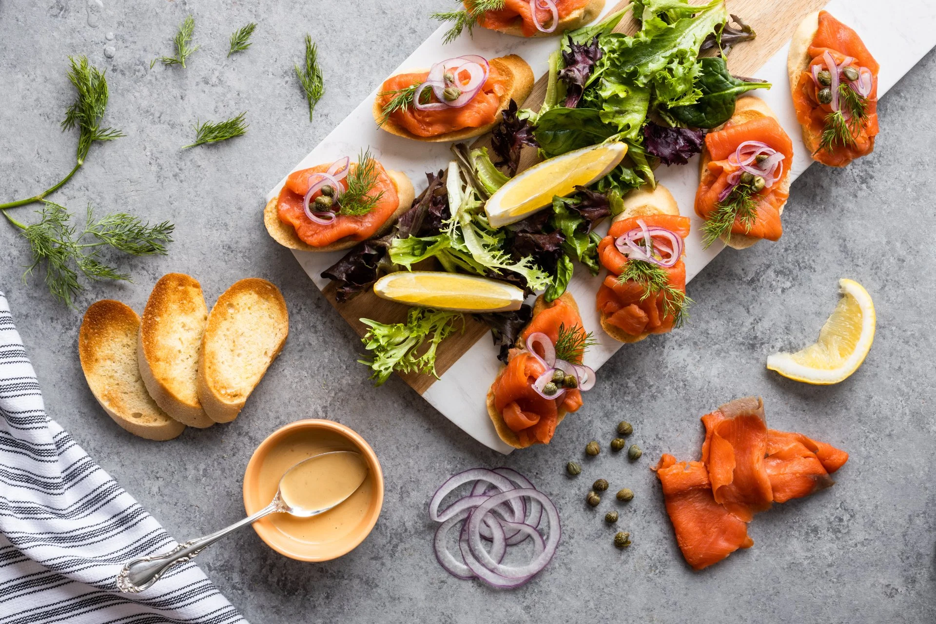 Smoked salmon and dill on crostini with capers, alongside a mixed green salad with lemon wedges, sliced onions, and a bowl of creamy sauce, on a stone surface.