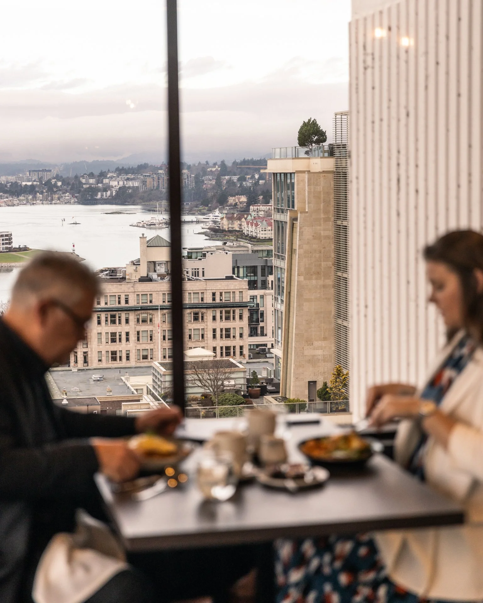 People eating breakfast at a table with a view of city buildings and a waterfront through large windows.
