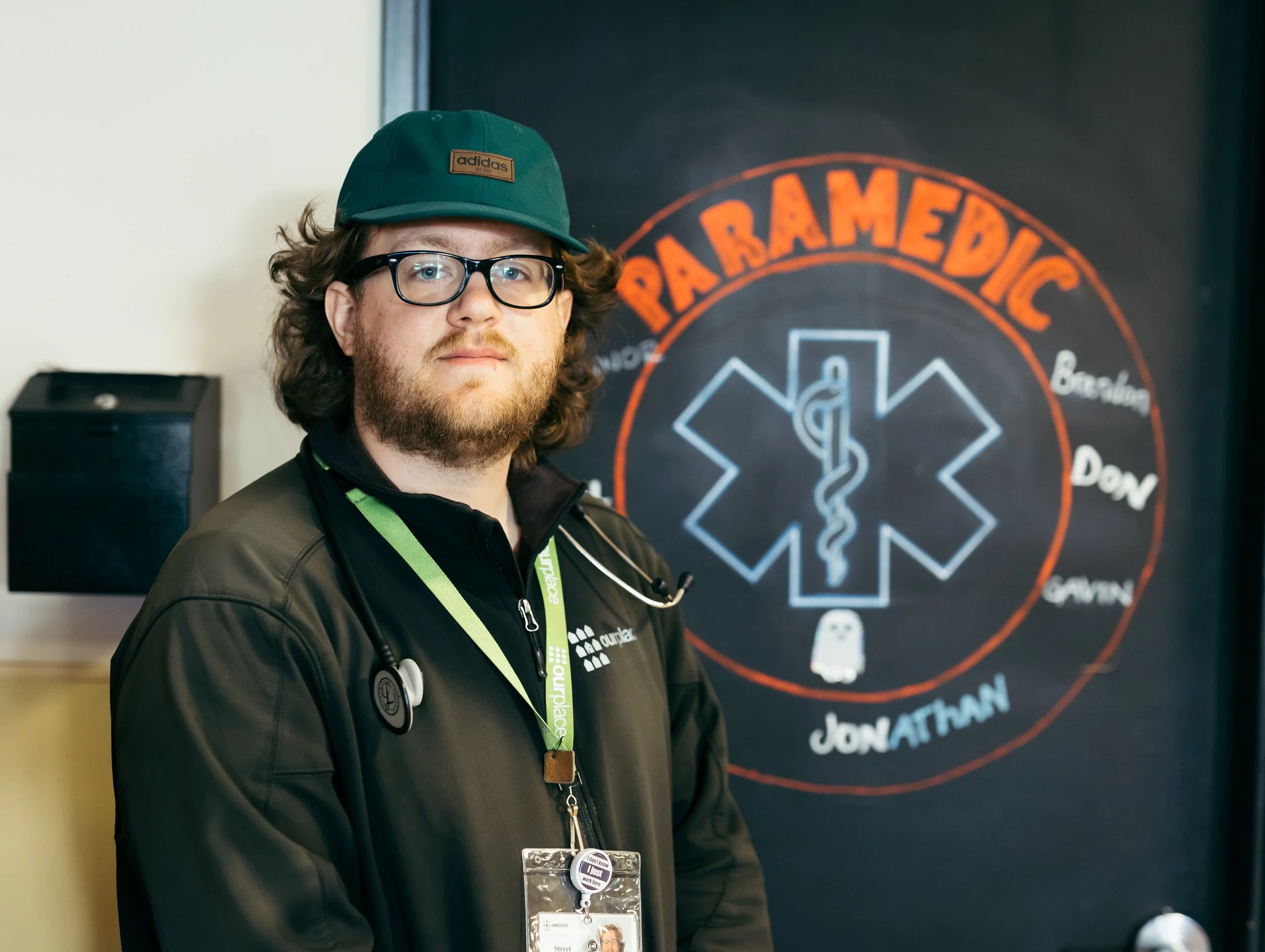 A man with curly hair, glasses, and a beard wearing a green hat and black jacket stands in front of a blackboard with orange and white chalk writing, including the word 'PARAMEDIC' and a medical symbol.