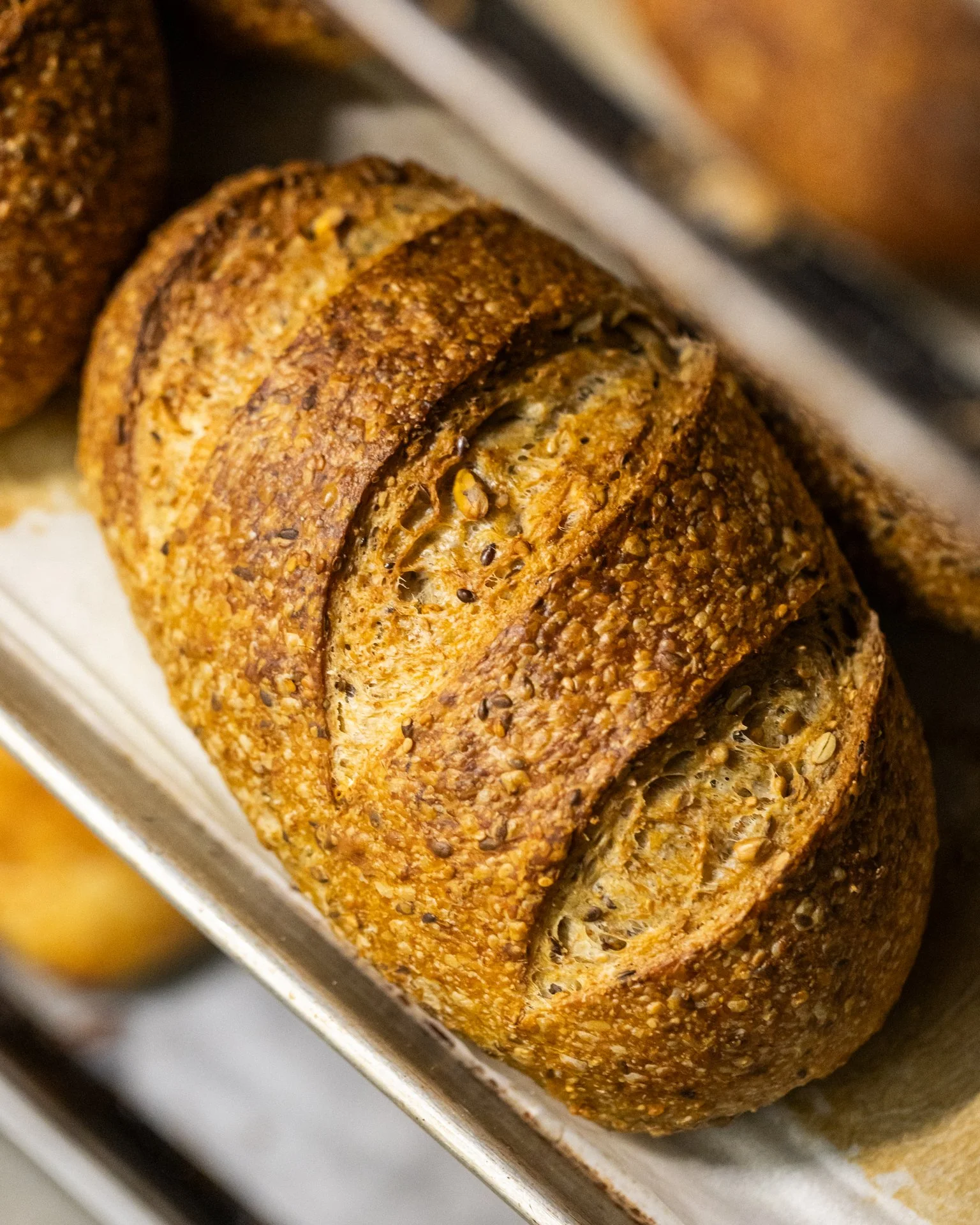 Close-up of a baked loaf of bread with a crust covered in sesame seeds and herbs, resting in a baking pan.