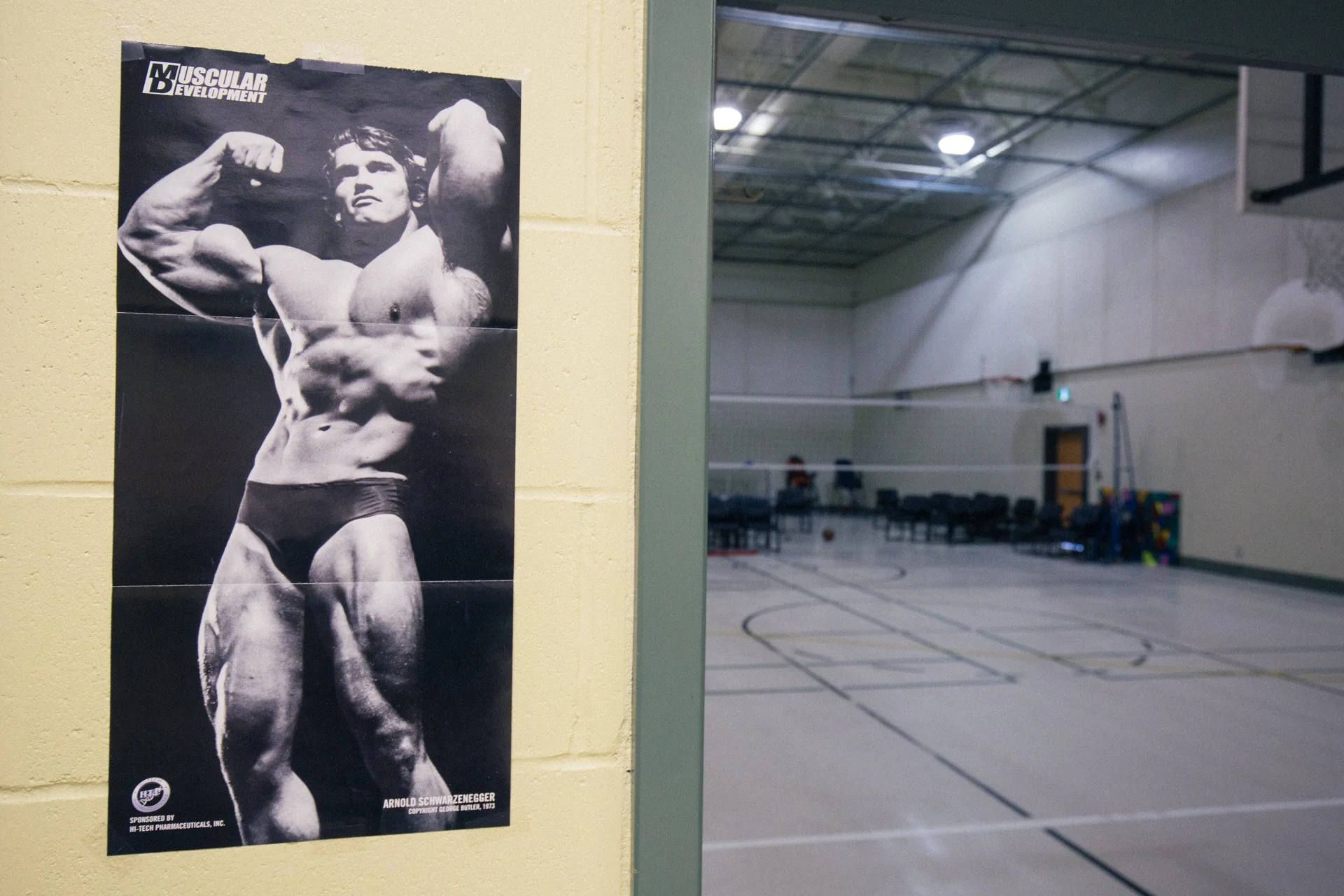 A black and white poster of Arnold Schwarzenegger flexing his arm in a muscle pose, displayed on a yellow wall inside a gymnasium.