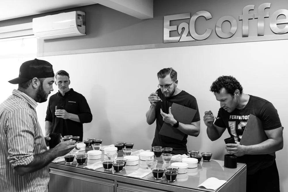 Four men sampling coffee at a tasting event in a modern room with a sign that reads 'E2 Coffee' on the wall.