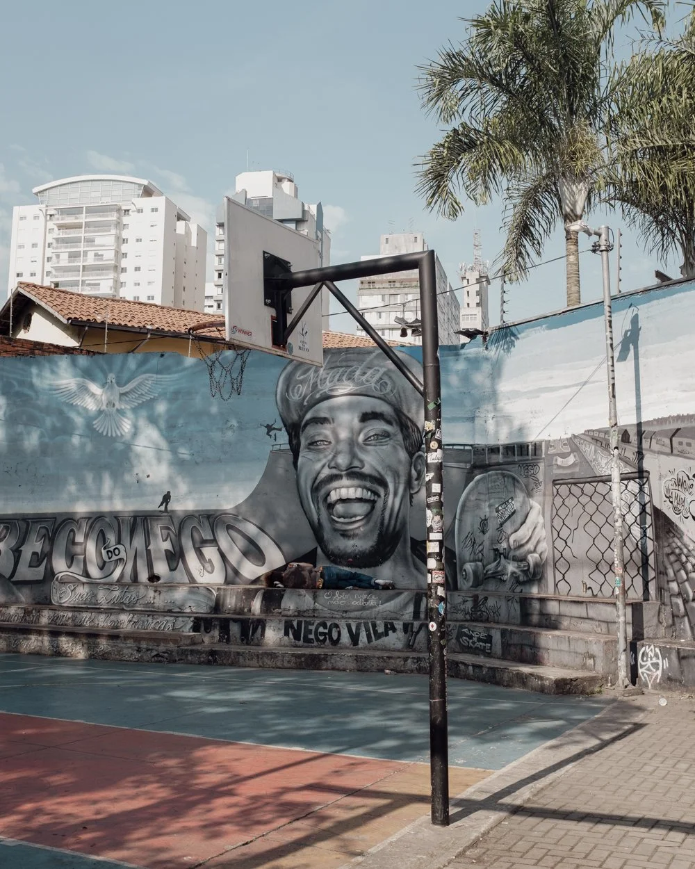 An outdoor basketball court with a mural of a smiling man holding a soda can, surrounded by graffiti, with a basketball hoop, palm trees, and high-rise buildings in the background.