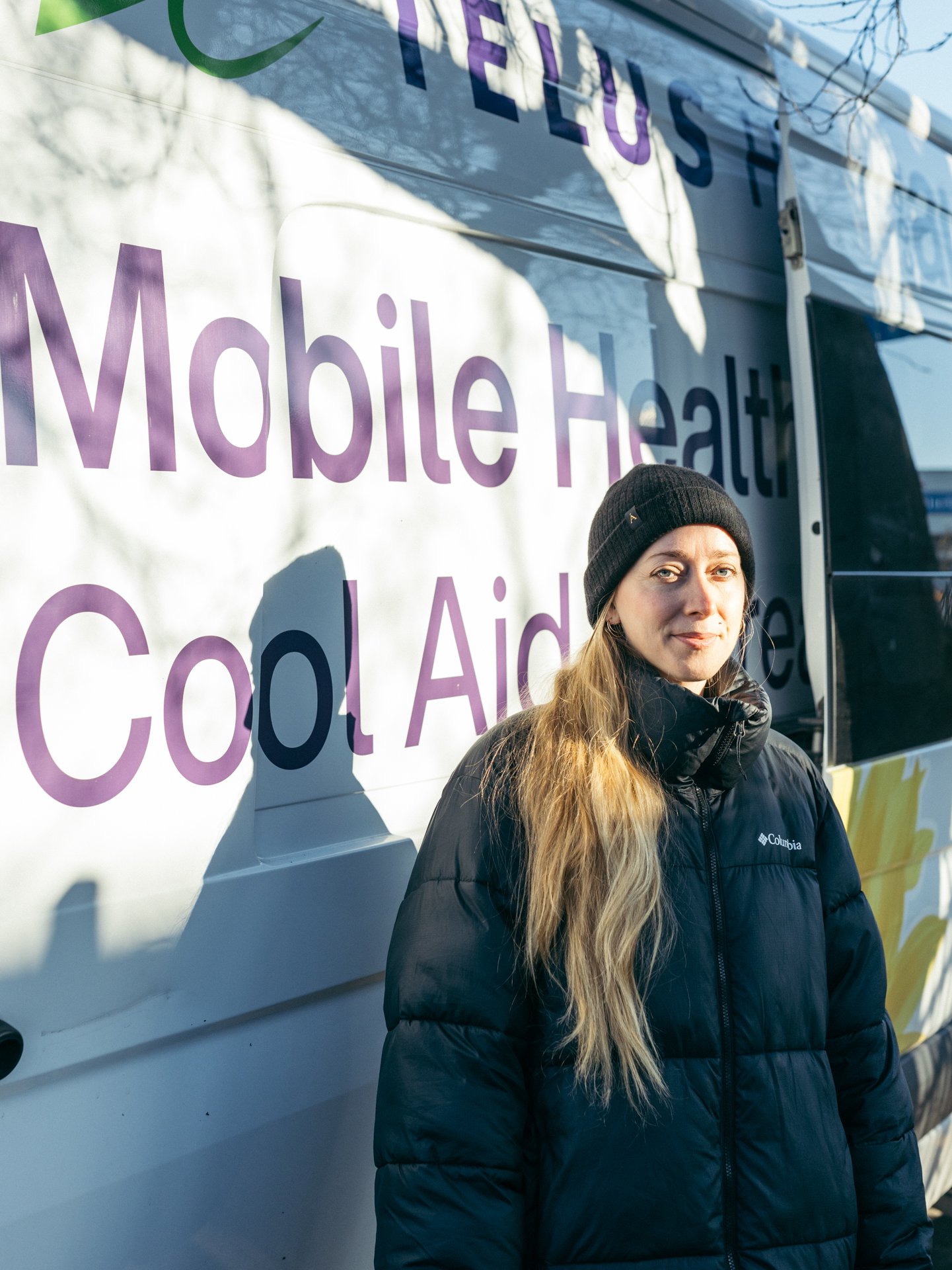 A young woman with long blonde hair wearing a black beanie and a black Columbia jacket standing outdoors in front of a Mobile Health vehicle with purple and green lettering. Shadows of tree branches are cast on the vehicle.