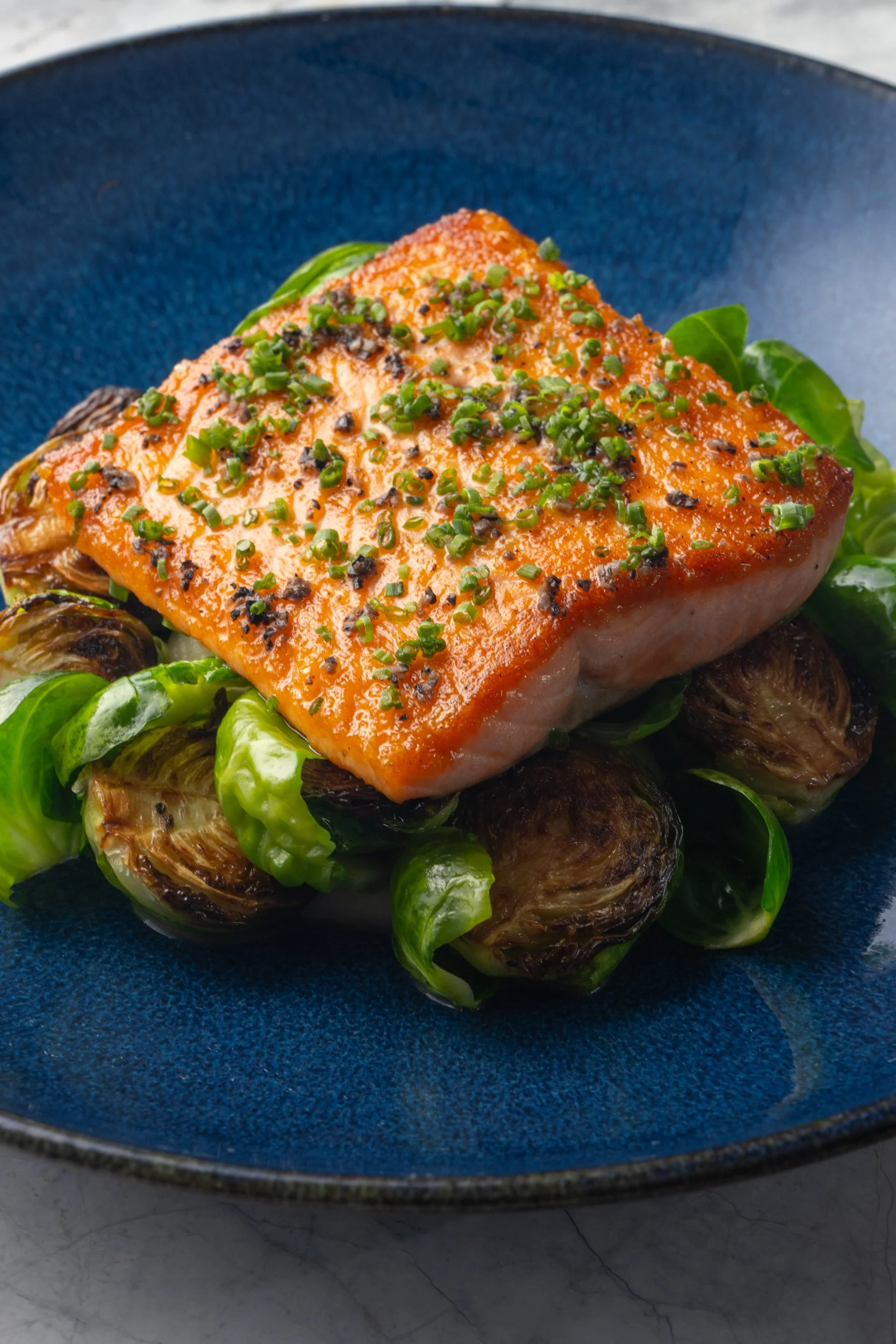 A cooked salmon fillet topped with chopped chives on a bed of roasted Brussels sprouts and green peppers, served on a blue ceramic plate.
