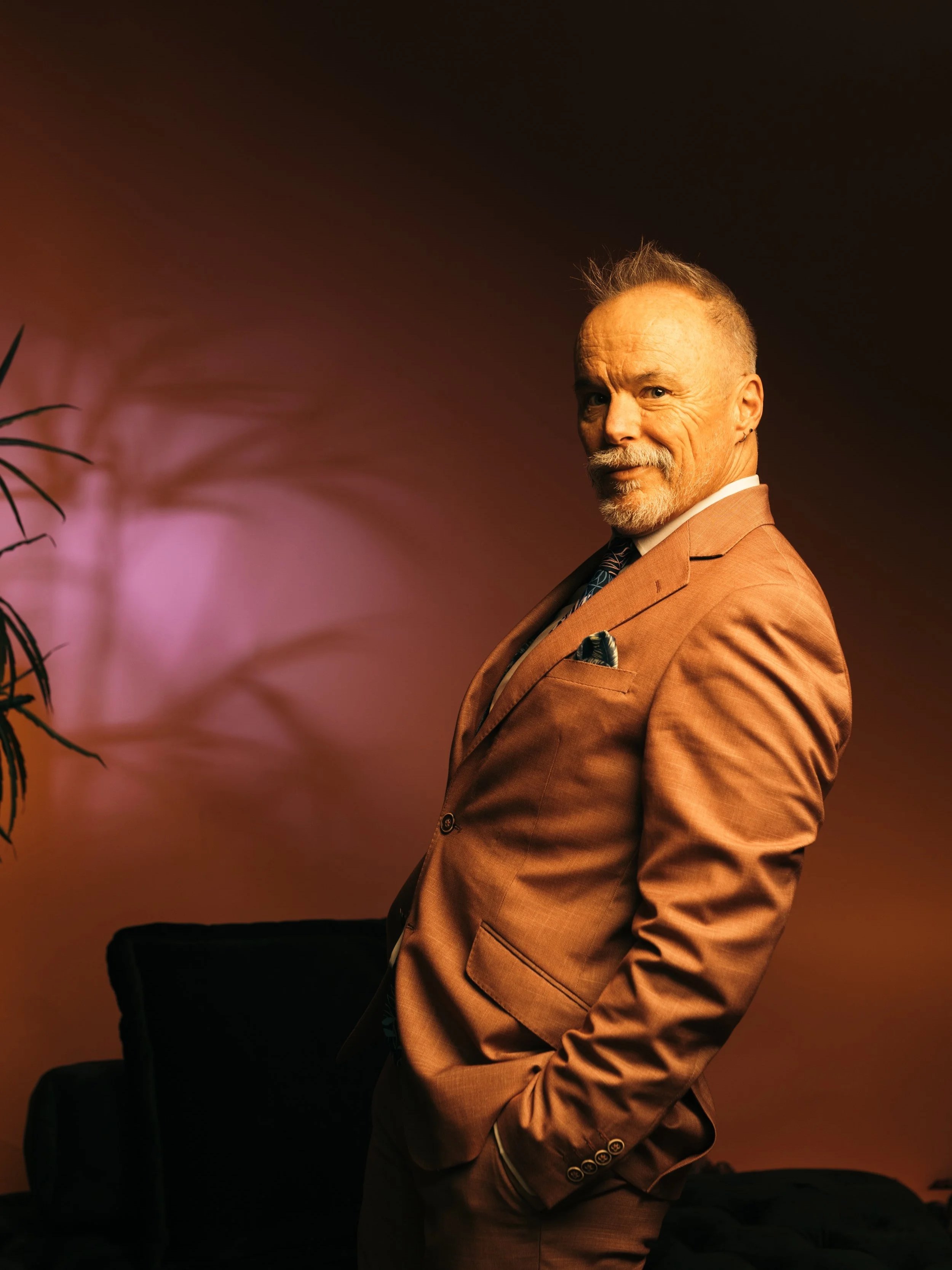 A man with a beard, wearing a brown suit and a patterned tie, poses in front of a dark background with dim lighting.