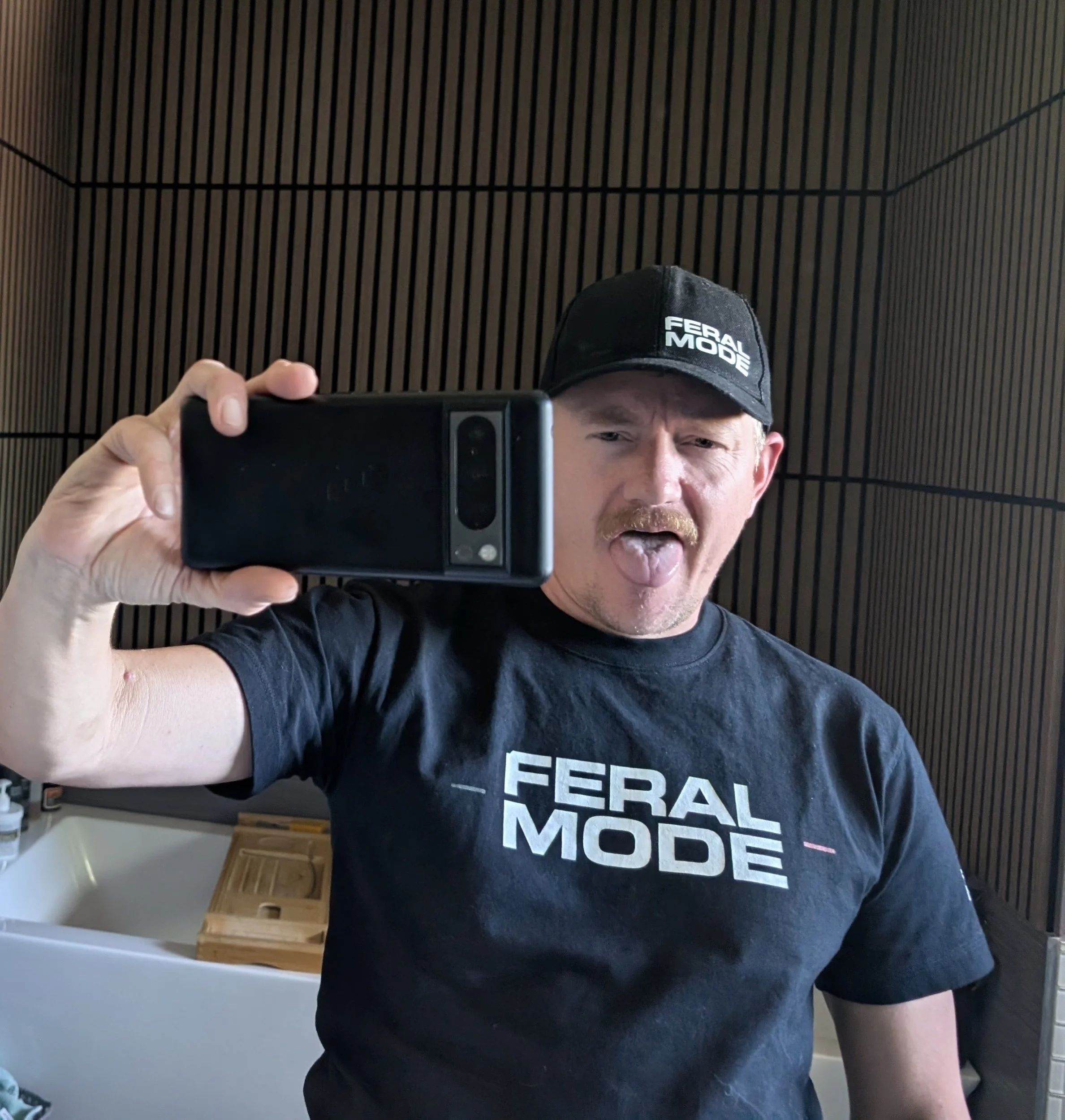 The founder takes a selfie in a mirror, wearing a black cap with white text reading 'Feral Mode' and a black T-shirt with white text. The background shows a wall with vertical wooden slats and some furniture.