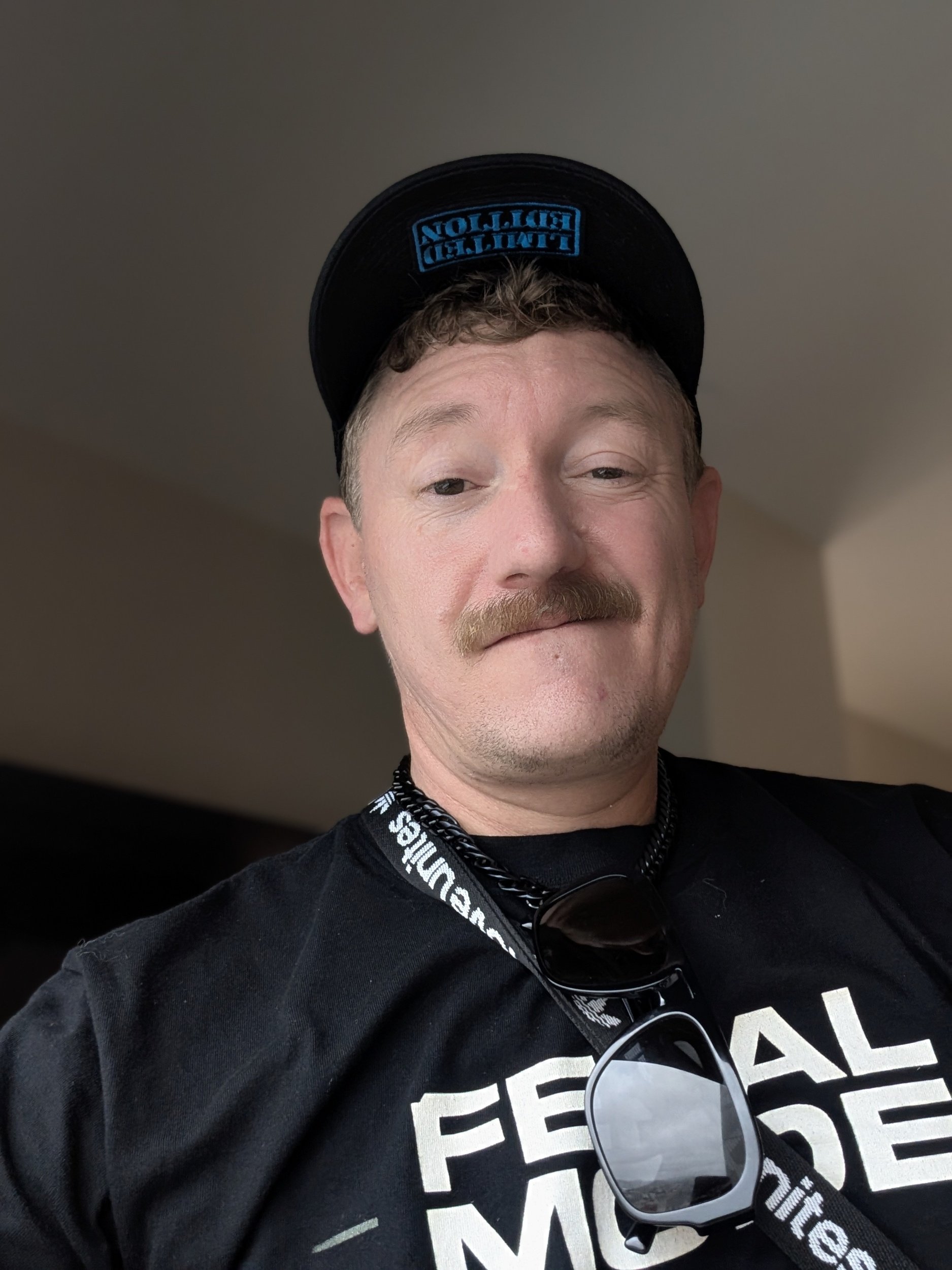 The founder wearing a black baseball cap with blue embroidered text and a black T-shirt with white text. He has short, light-colored hair, and sunglasses hanging from his shirt collar. The background is an indoor setting with a plain wall.