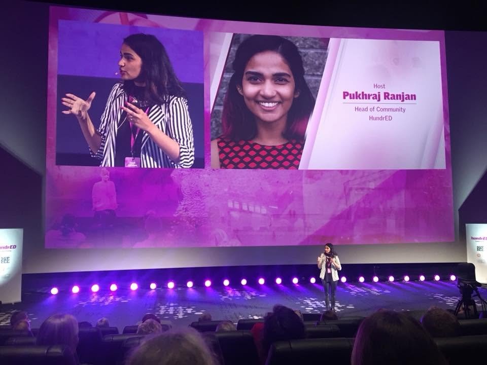A woman speaking on stage at a conference, with a large screen behind her displaying images of another woman and event information, including her name Pukhraj Ranjan and her role as Head of Community at HundrED.