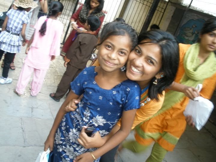 A girl in a blue dress smiling with a woman in an orange traditional dress, surrounded by other children and adults in a busy outdoor setting.