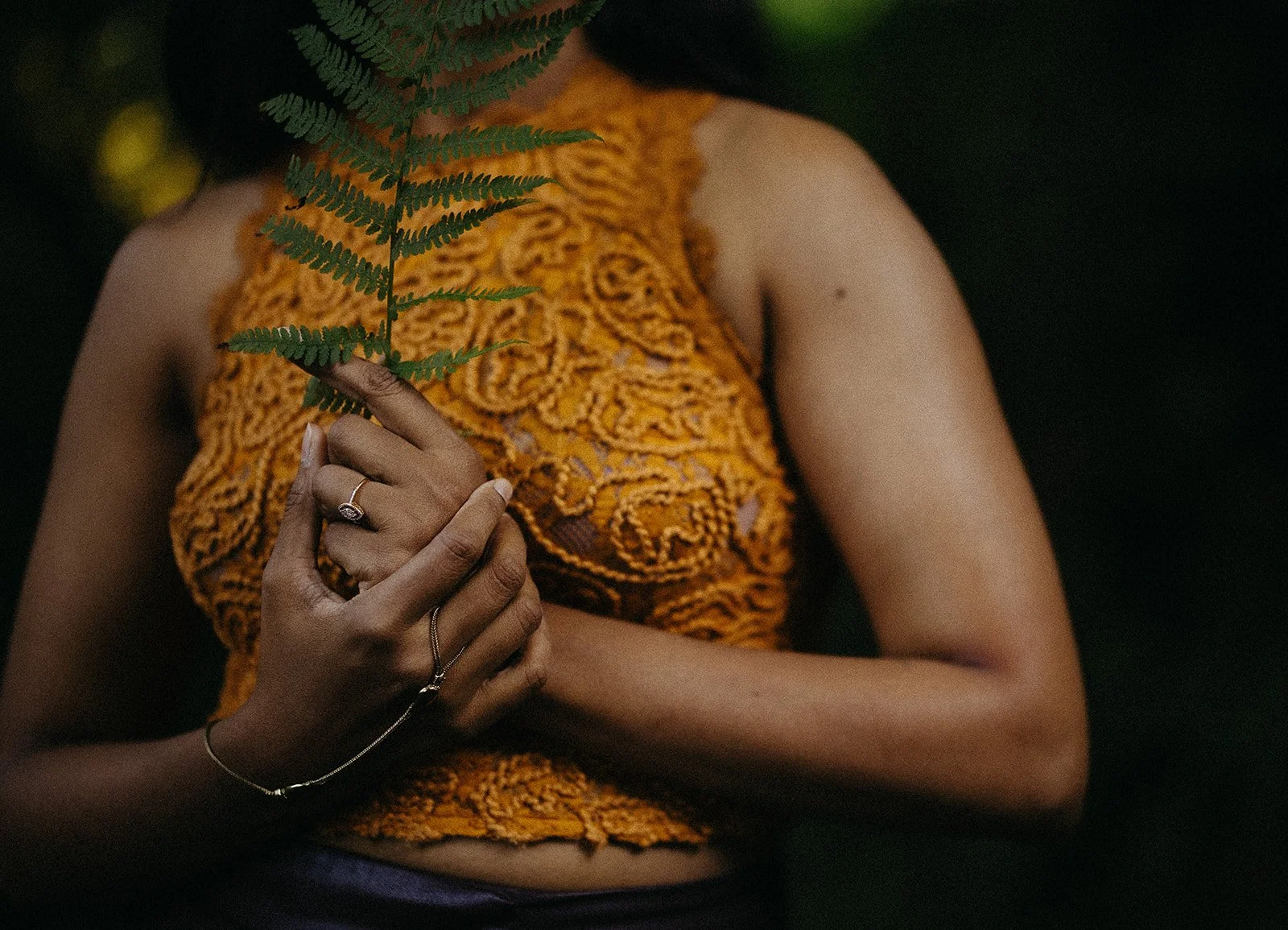 A woman wearing a colorful orange lace top, holding a fern leaf in front of her face, with dark background.