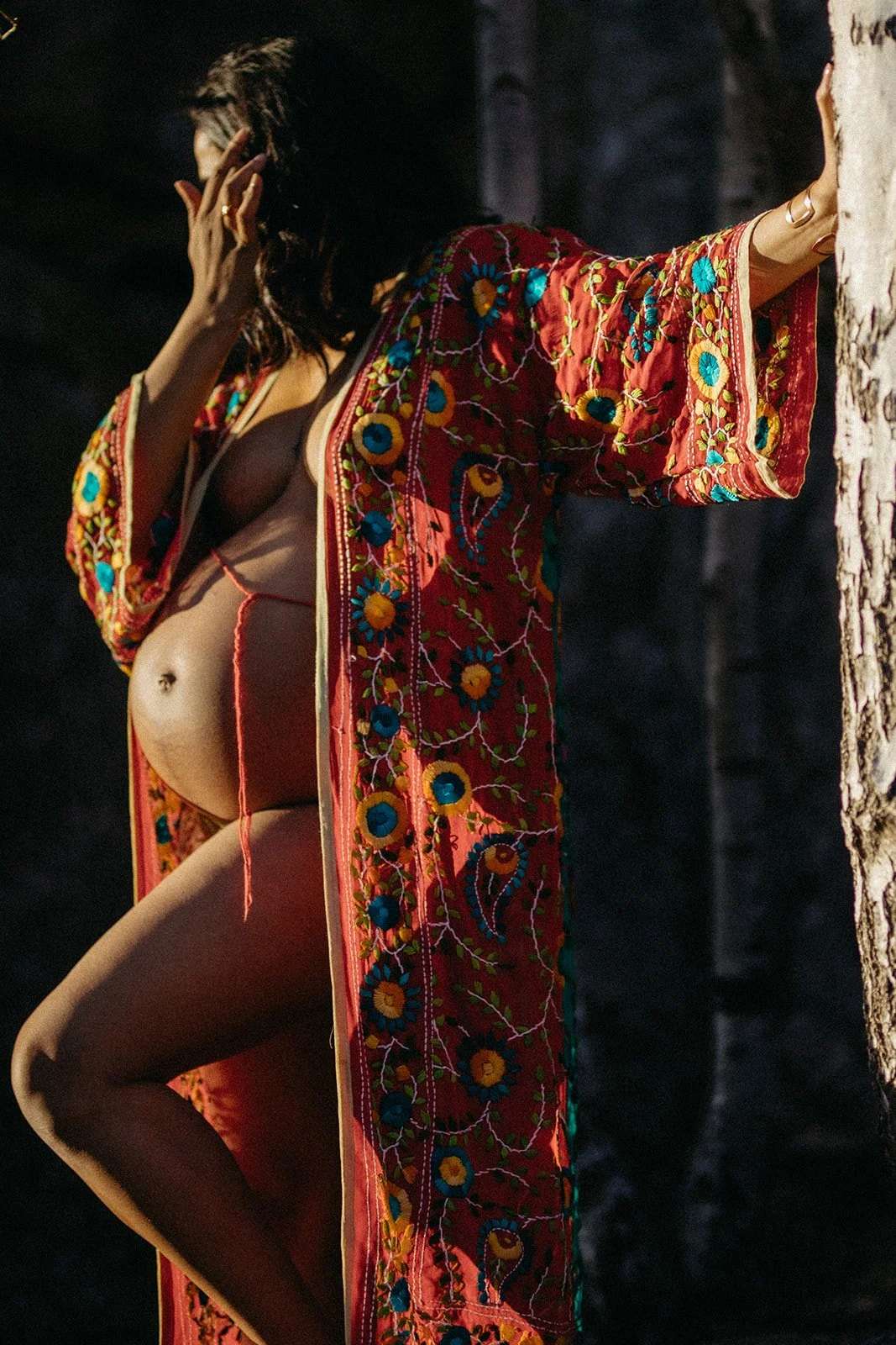 A woman wearing a sheer, embroidered kimono with floral patterns, leaning against a tree in a dark setting.