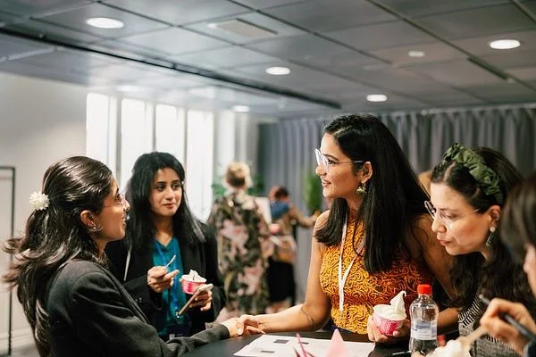 Five women, including one in a yellow top, are engaging in conversation at a table in a well-lit room with large windows and gray curtains. One woman is holding a cupcake, while another has a water bottle.