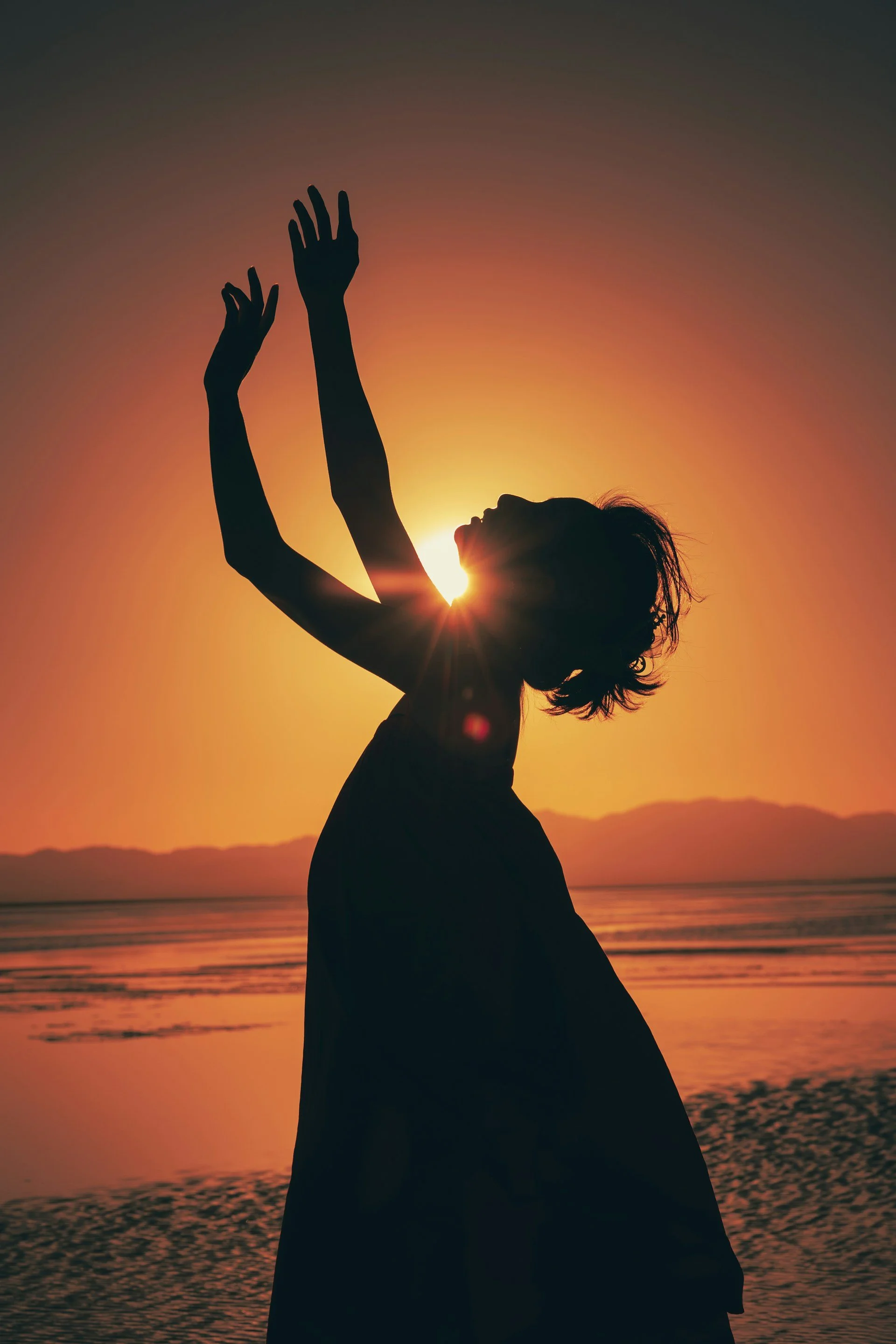 Silhouette of a woman with arms raised, standing on a beach at sunset, with mountains and water in the background.