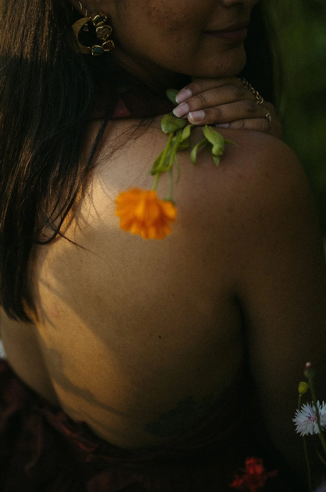 Close-up of a woman with dark hair, wearing gold earrings, holding a yellow flower near her shoulder, with a blurred background.